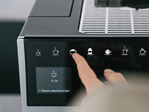 Bir parmak, kahve makinesinin siyah kontrol panelinde bir kahve fincanı simgesine basıyor. Metin 'Lütfen içeceği seçin' diyor.