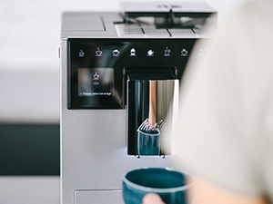 Ekranında kahve fincanı simgesi olan modern bir kahve makinesi. Bir kişi mavi bir fincan tutuyor.