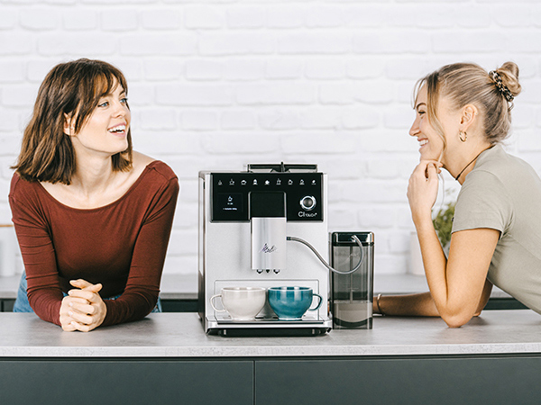 İki kadın kahve makinesinin yanında birbirlerine gülümsüyor. Arka planda beyaz tuğla duvar. Fincanlar hazır.