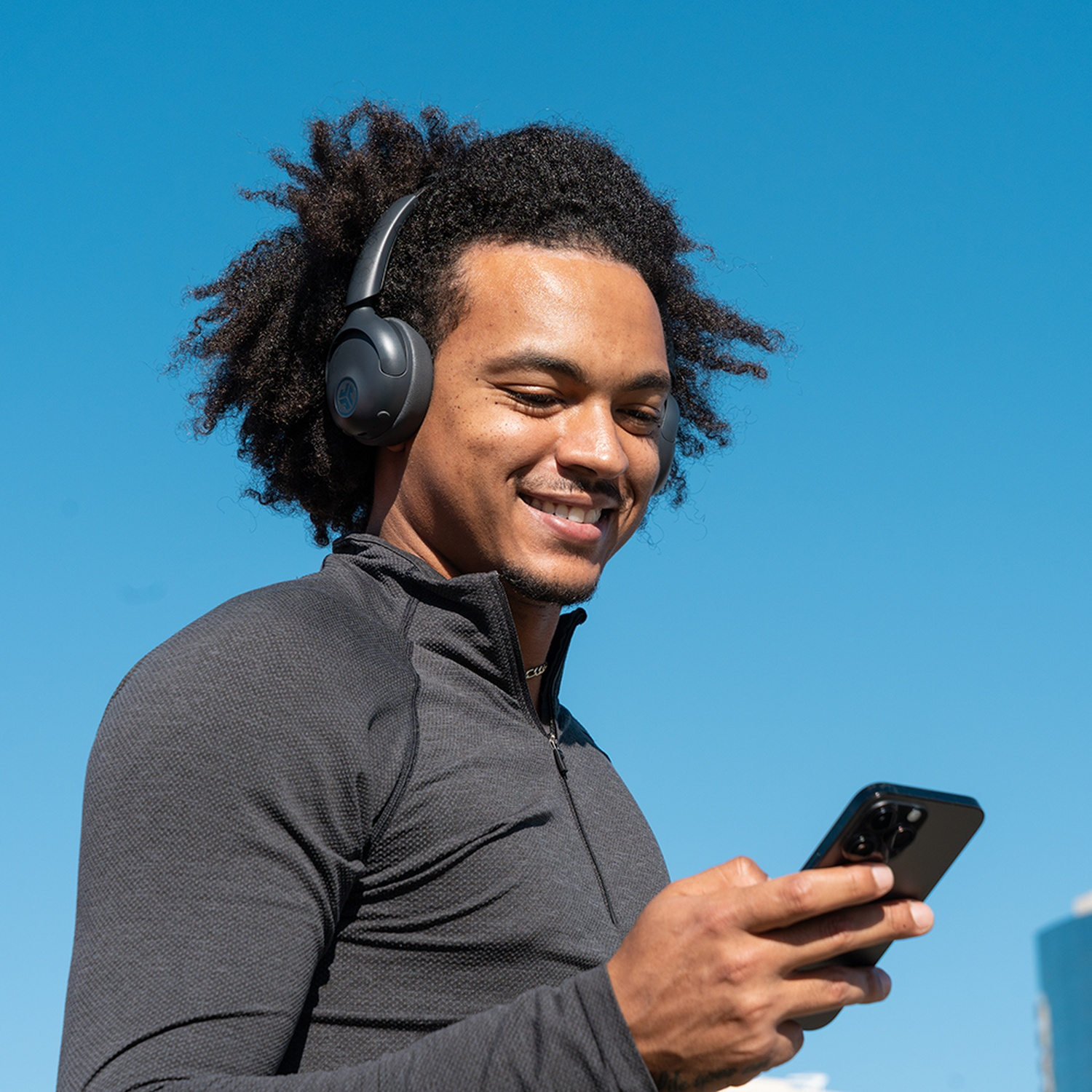 Un homme souriant avec un casque regarde son téléphone. Il porte une veste grise contre un ciel bleu vif.