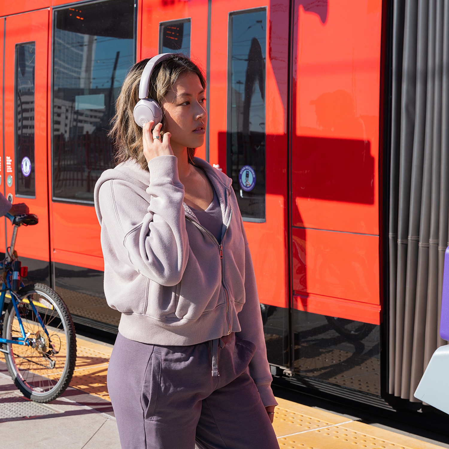Une femme avec un casque se tient près d'un train rouge, touchant une oreille. Un vélo et une plateforme sont également visibles.