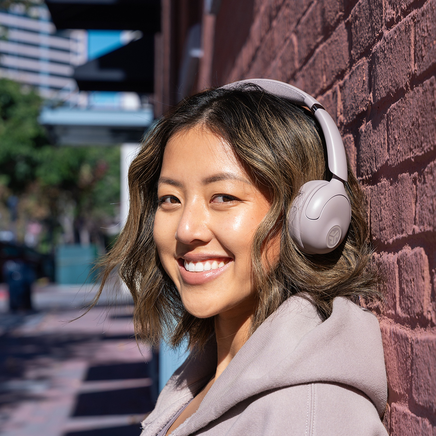 Une femme asiatique souriante avec un casque audio s'appuie contre un mur de briques. Elle porte un sweat à capuche. Des bâtiments sont en arrière-plan.