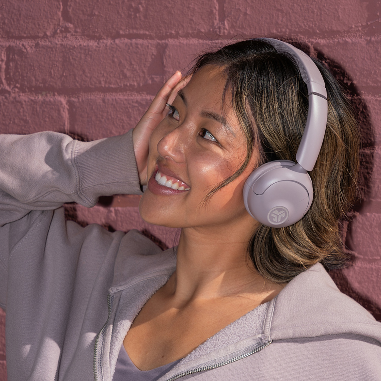 Une femme sourit, portant un casque et un sweat à capuche zippé, appuyée contre un mur de briques. Elle se touche la tête.