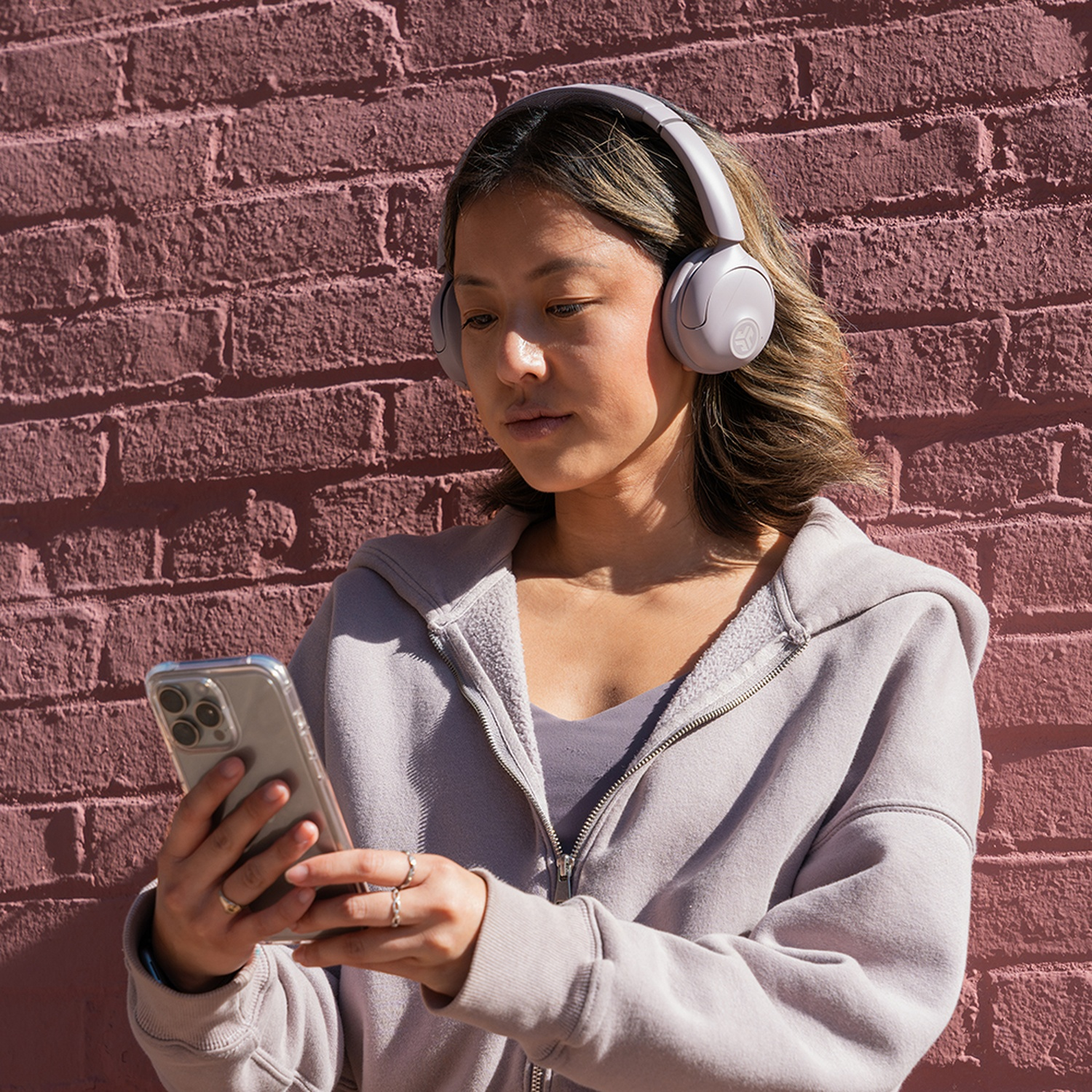 Une femme portant un casque et un sweat à capuche regarde son téléphone. Elle se tient devant un mur de briques rouges, à l'extérieur.
