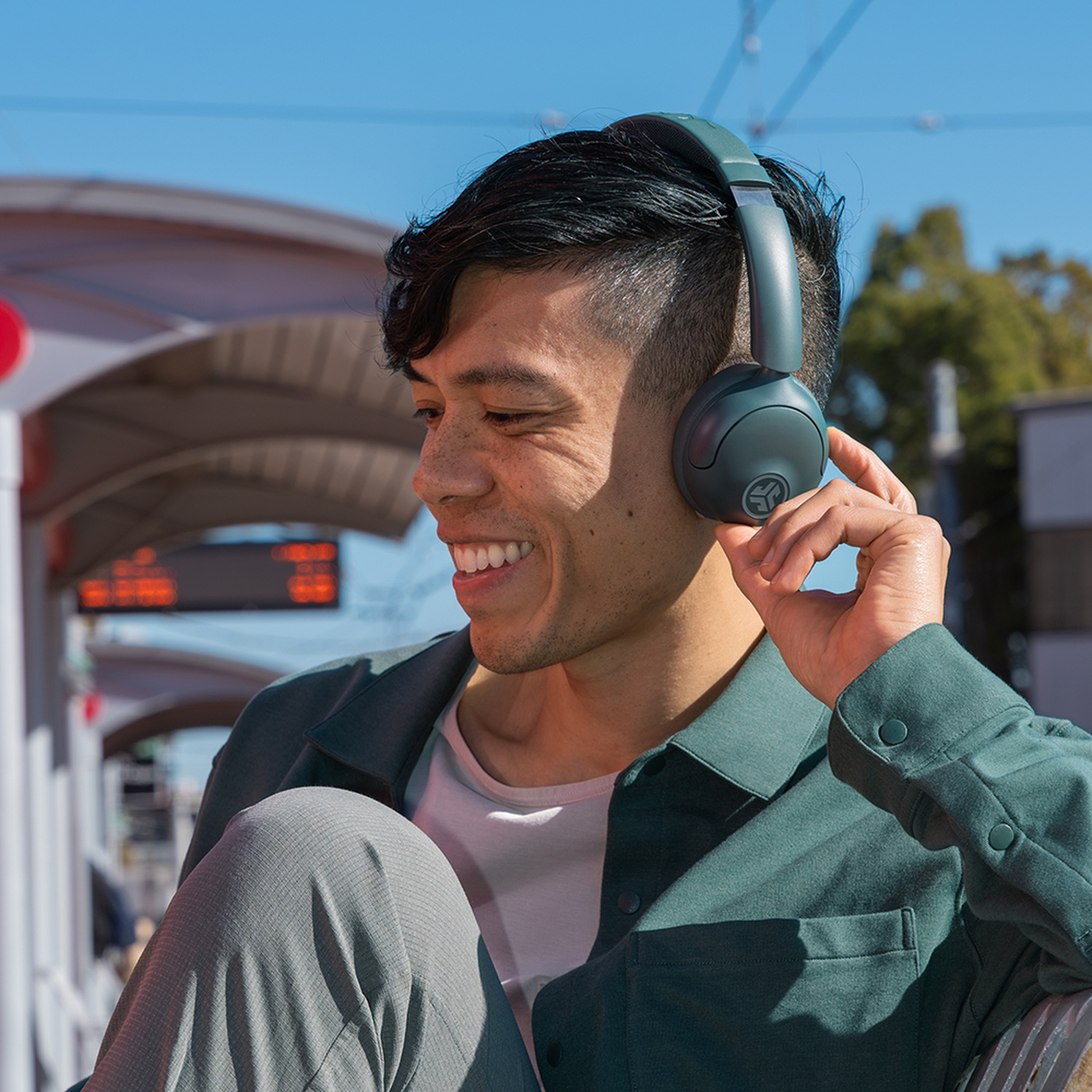 Un homme avec un casque sourit dans une gare. Il porte une chemise verte et un pantalon gris. Le ciel est bleu.