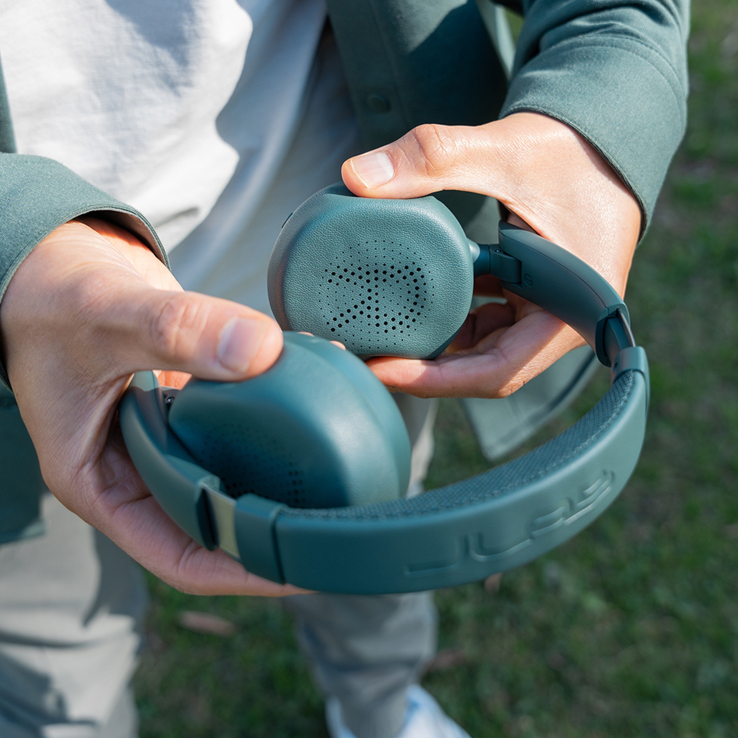 Une personne tient un casque vert d'eau à l'extérieur. Le casque est ouvert, montrant les écouteurs et l'arceau. De l'herbe verte en arrière-plan.