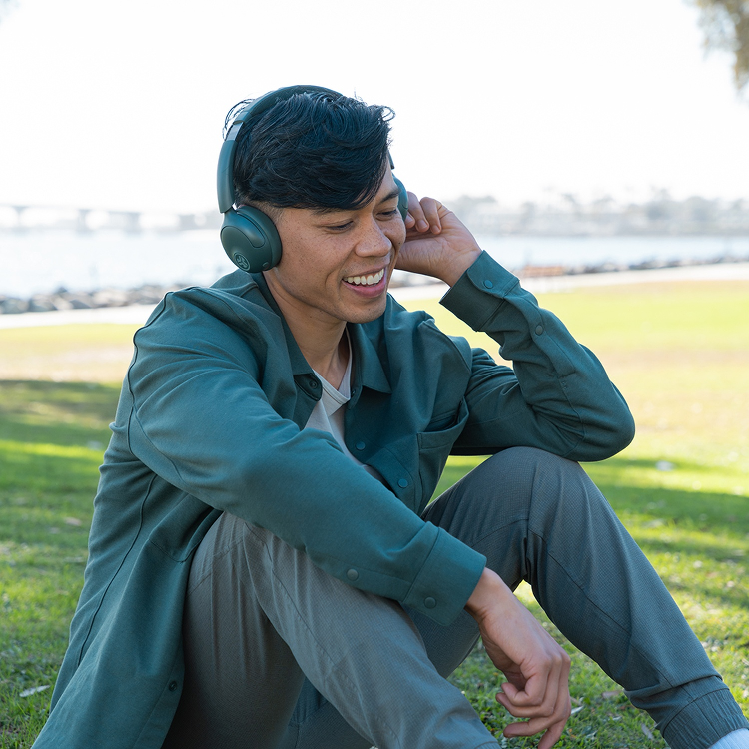 Un homme souriant portant un casque est assis sur l'herbe. Il porte une chemise et un pantalon verts. Un pont et de l'eau en arrière-plan.