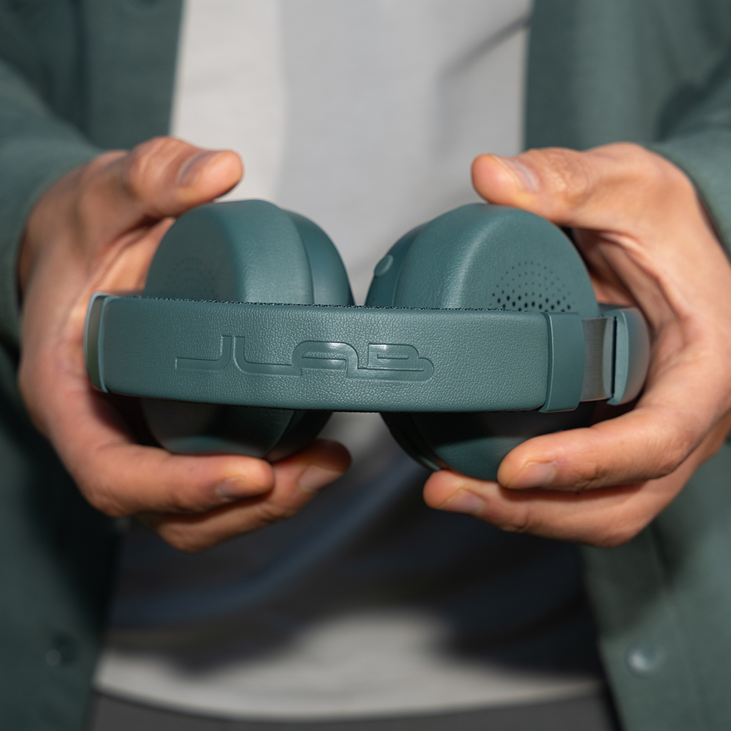 Personne tenant un casque de couleur teal avec le logo JLab. La personne porte une veste verte et une chemise blanche.