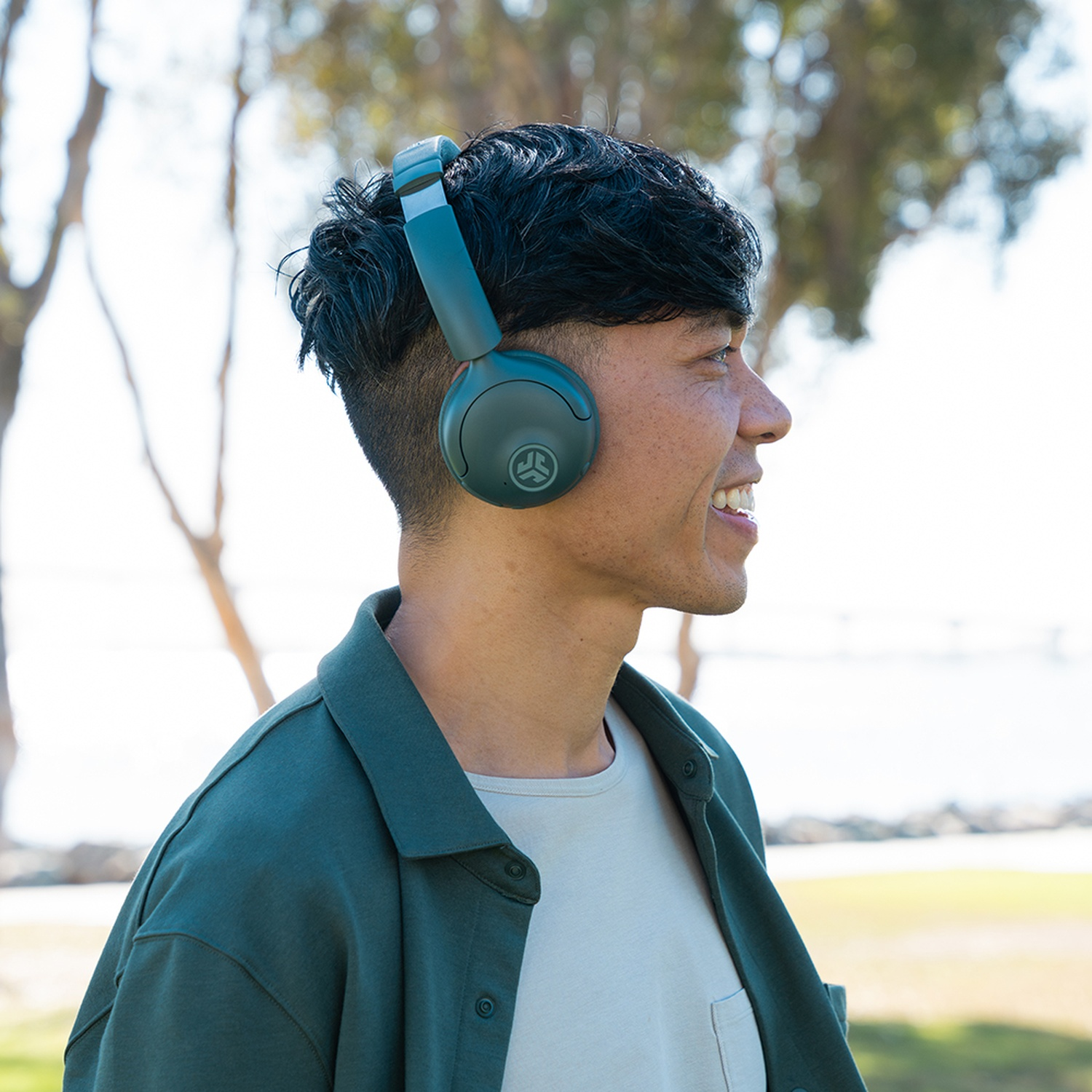 Un homme portant un casque audio sourit, regardant vers la droite. Il porte une veste verte et une chemise claire. Des arbres et un fond ensoleillé sont visibles.