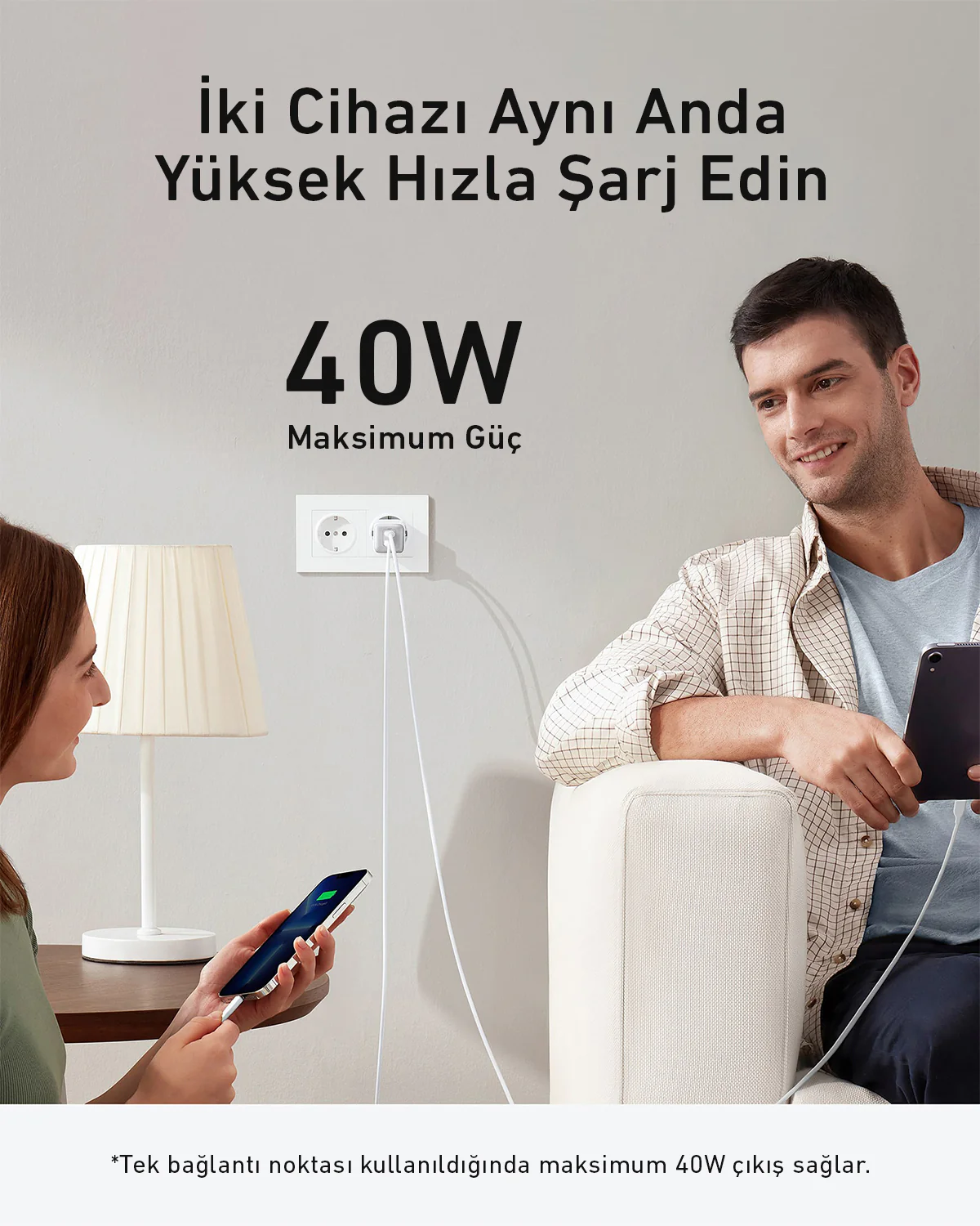 Bir erkek ve bir kadın, bir duvar prizine takılı bir şarj cihazıyla cihazları şarj ediyor. 40W metni görünür. Ayrıca bir lamba ve bir kanepe de var.