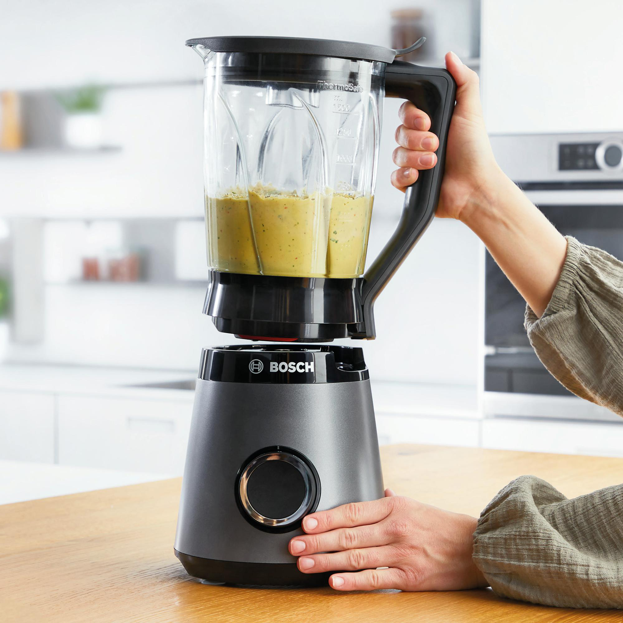 Eine Person hält einen grauen Bosch-Mixer mit grüner Flüssigkeit. Der Mixer steht auf einer Holzoberfläche.
