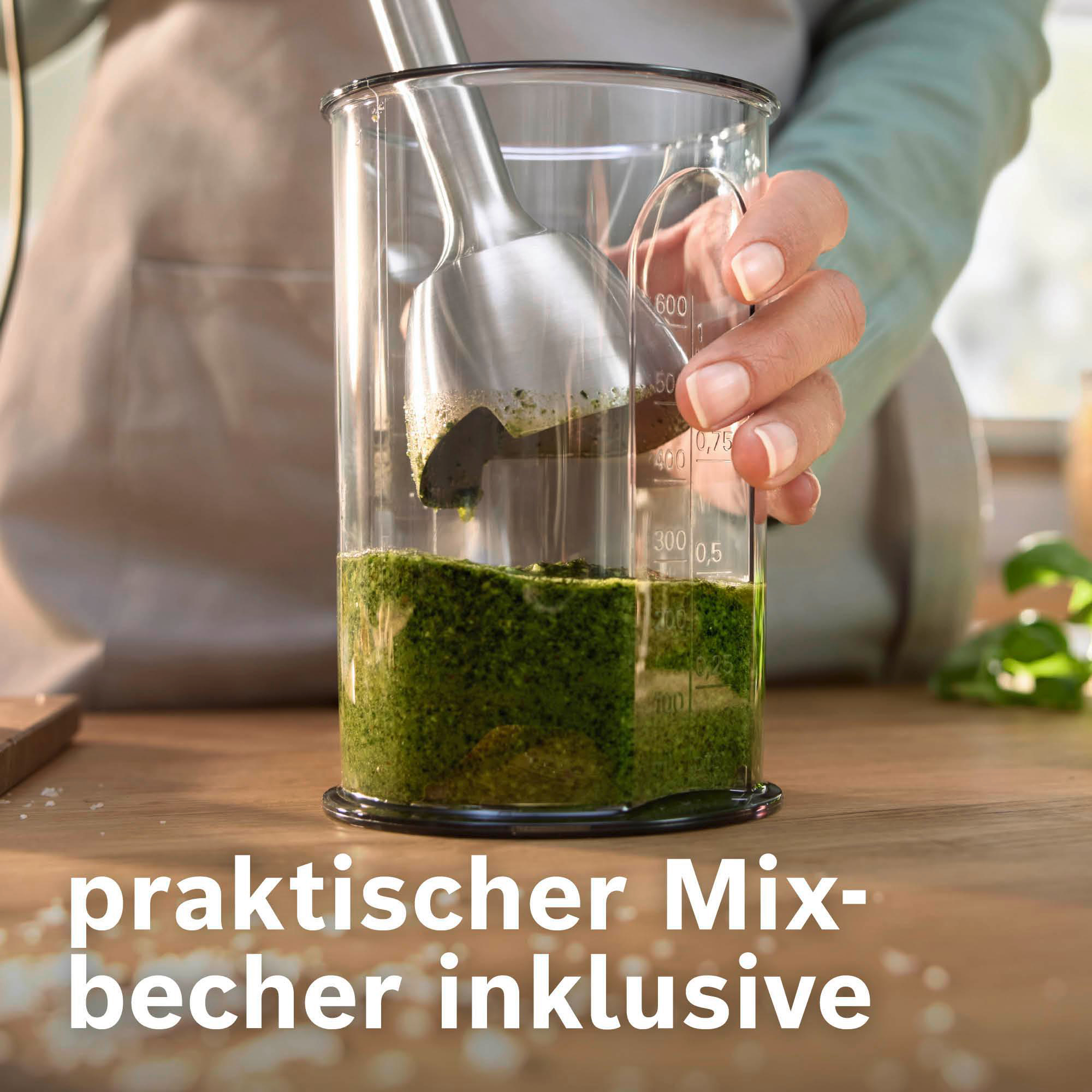 Eine Hand hält einen Mixer in einem Glas. Das Glas enthält grüne Sauce. Der Hintergrund ist ein Holztisch.