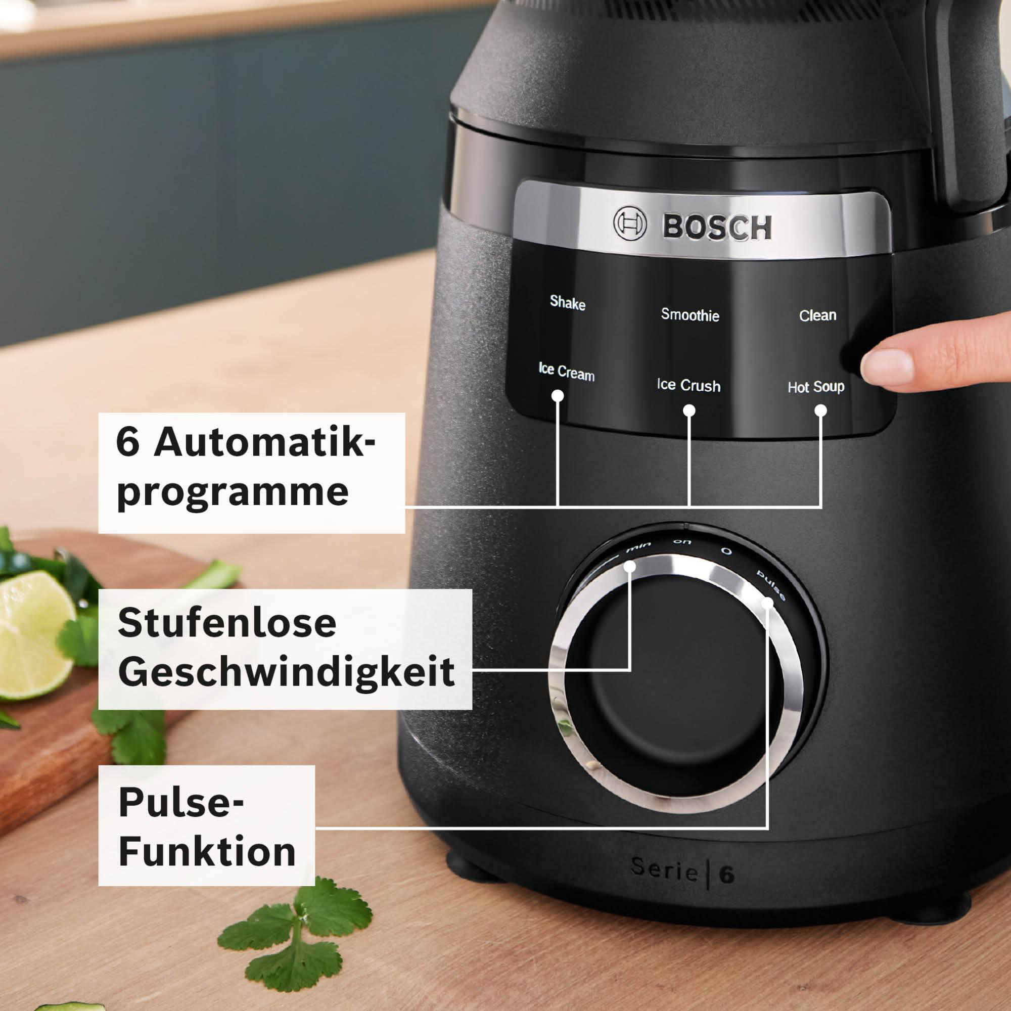 Ein schwarzer Bosch-Mixer mit Bedienfeld, eine Hand berührt eine Taste und Textbeschriftungen. Gemüse und Limette im Vordergrund.
