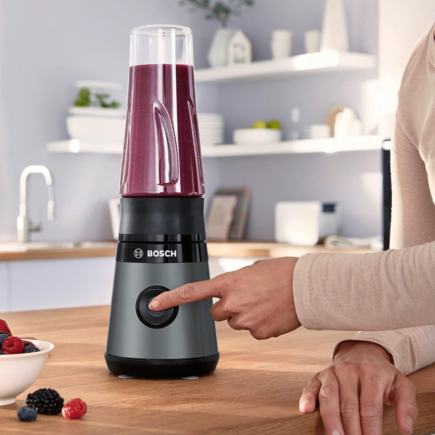 Eine Person drückt den Knopf eines grauen Bosch-Mixers mit einem lila Smoothie. Beeren liegen auf einem Holztisch.