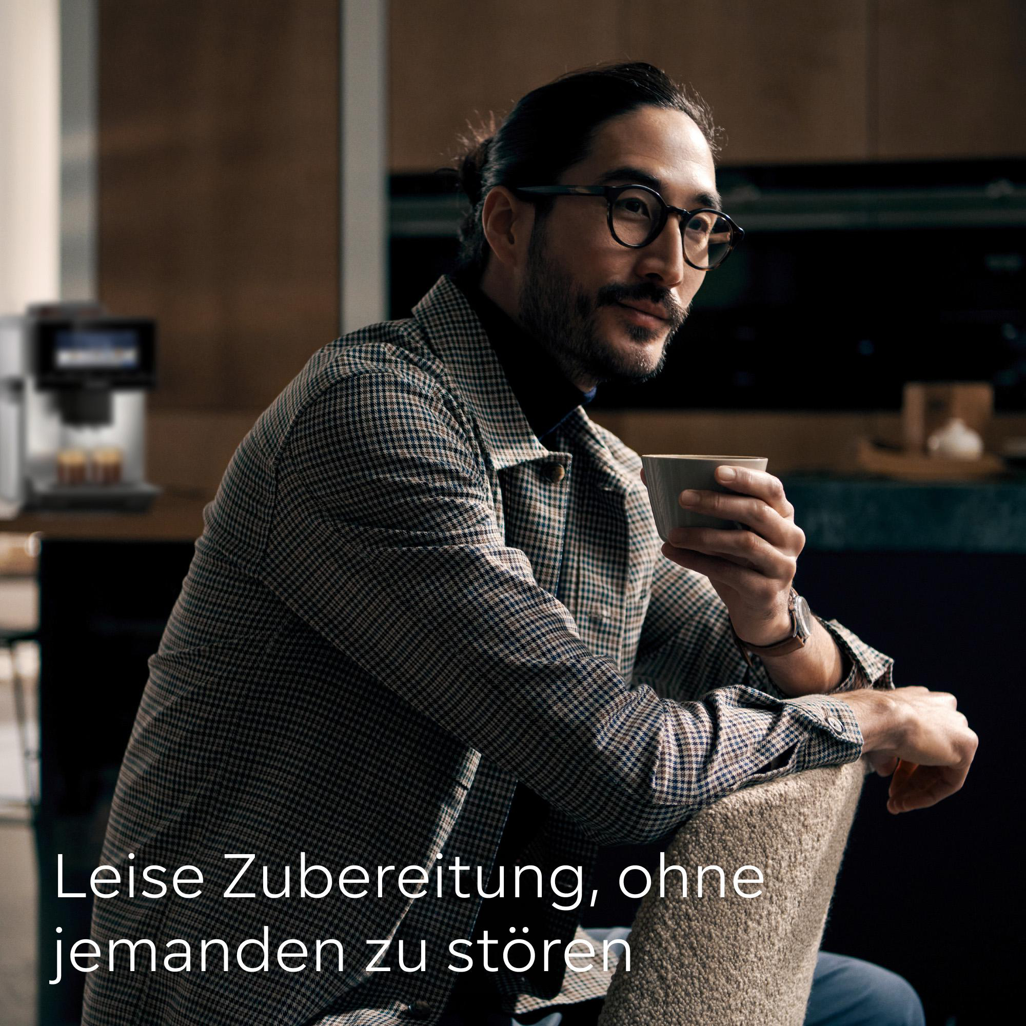 Ein Mann in einer karierten Jacke hält eine Tasse und sitzt in der Nähe einer Kaffeemaschine. Der Hintergrund ist verschwommen.