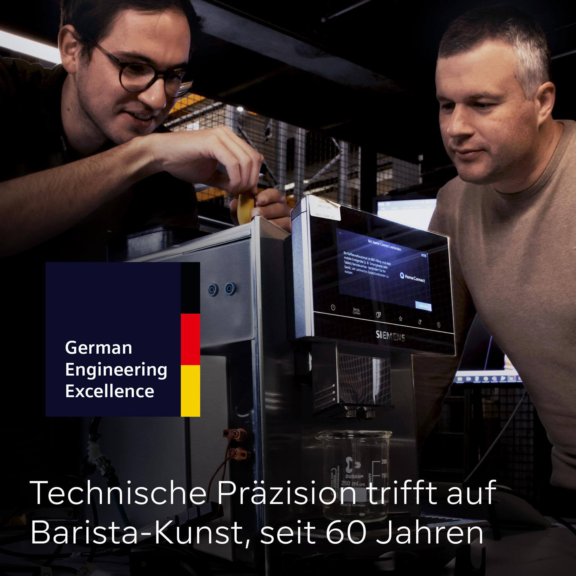 Zwei Männer an einer Kaffeemaschine. Einer justiert etwas, der andere schaut zu. Siemens-Logo sichtbar.