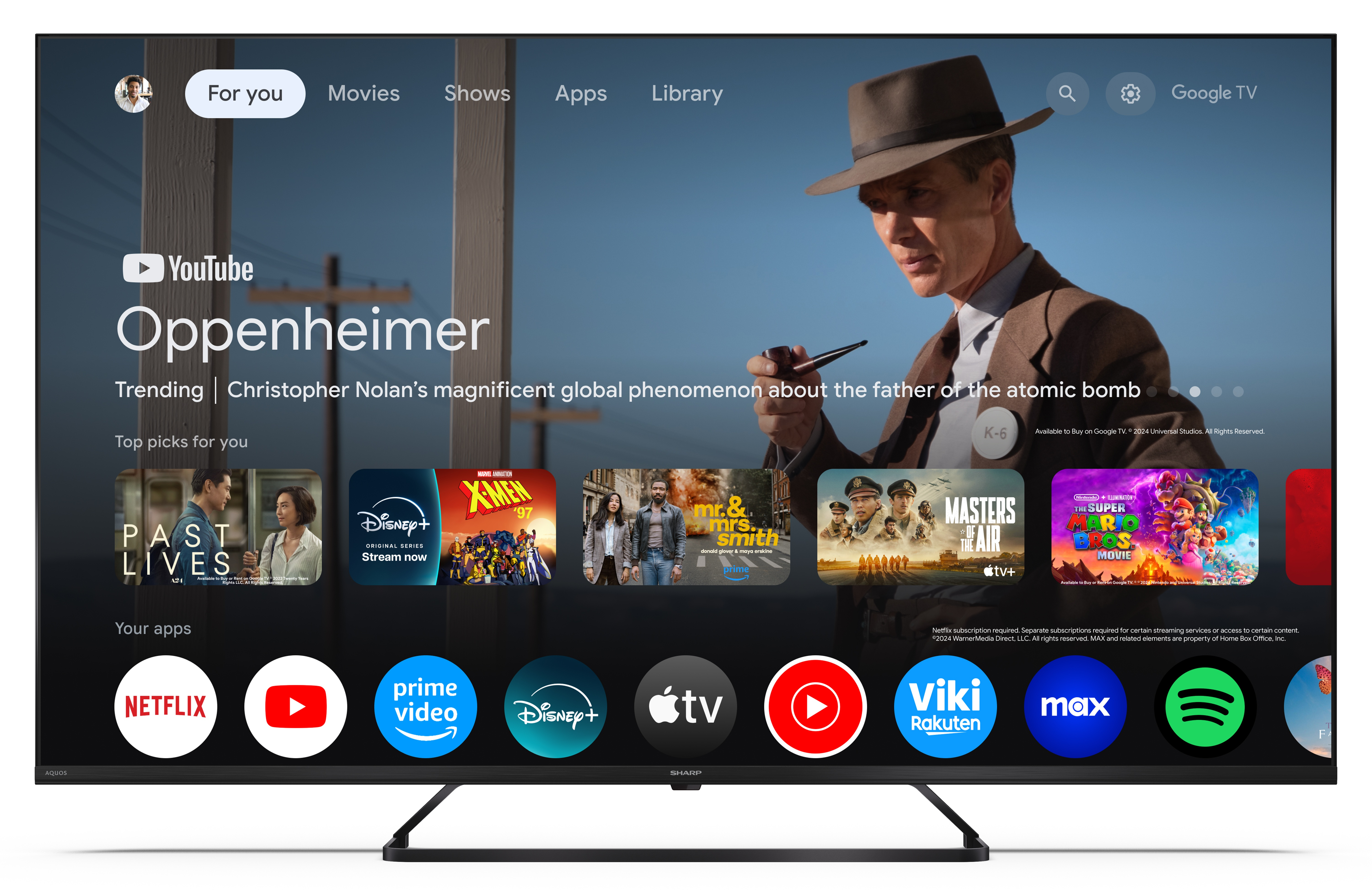 Ekran telewizora wyświetla interfejs Google TV z tytułami filmów i aplikacjami. Mężczyzna w kapeluszu jest w tle.