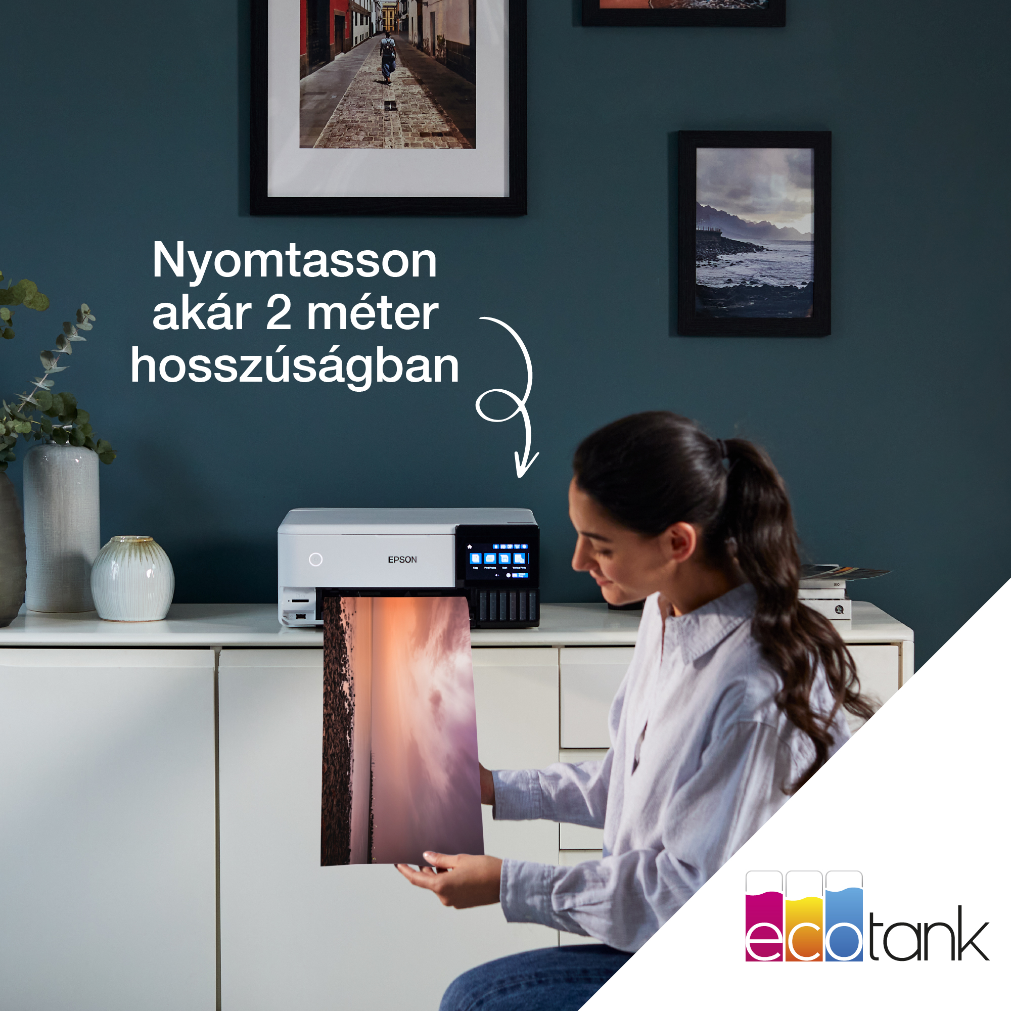 Egy nő nagy fotót nyomtat egy Epson nyomtatóval. Képek lógnak a falon. Fehér szekrény és vázák láthatók.