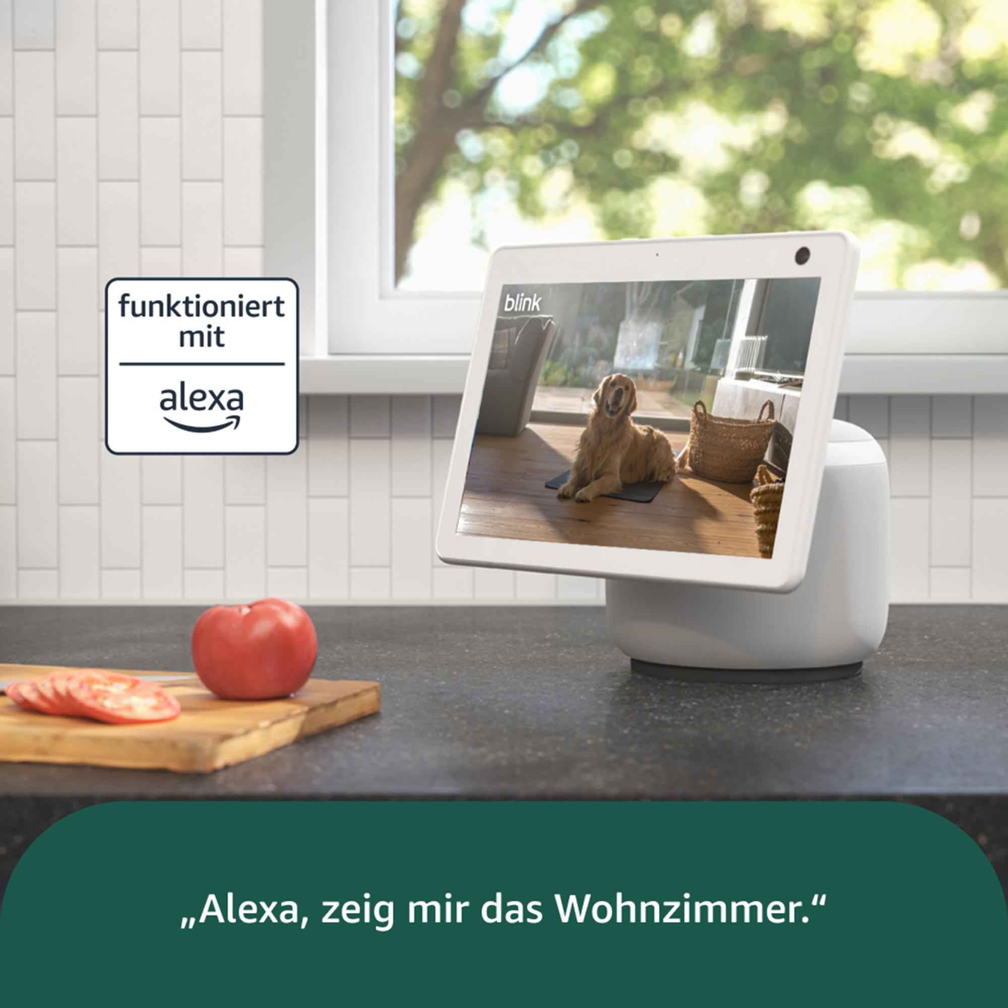 Ein Smart-Display zeigt einen Hund. Auf der Theke liegen eine Tomate und Tomatenscheiben. Auf dem Bildschirm steht: 'Alexa, zeig mir das Wohnzimmer'.