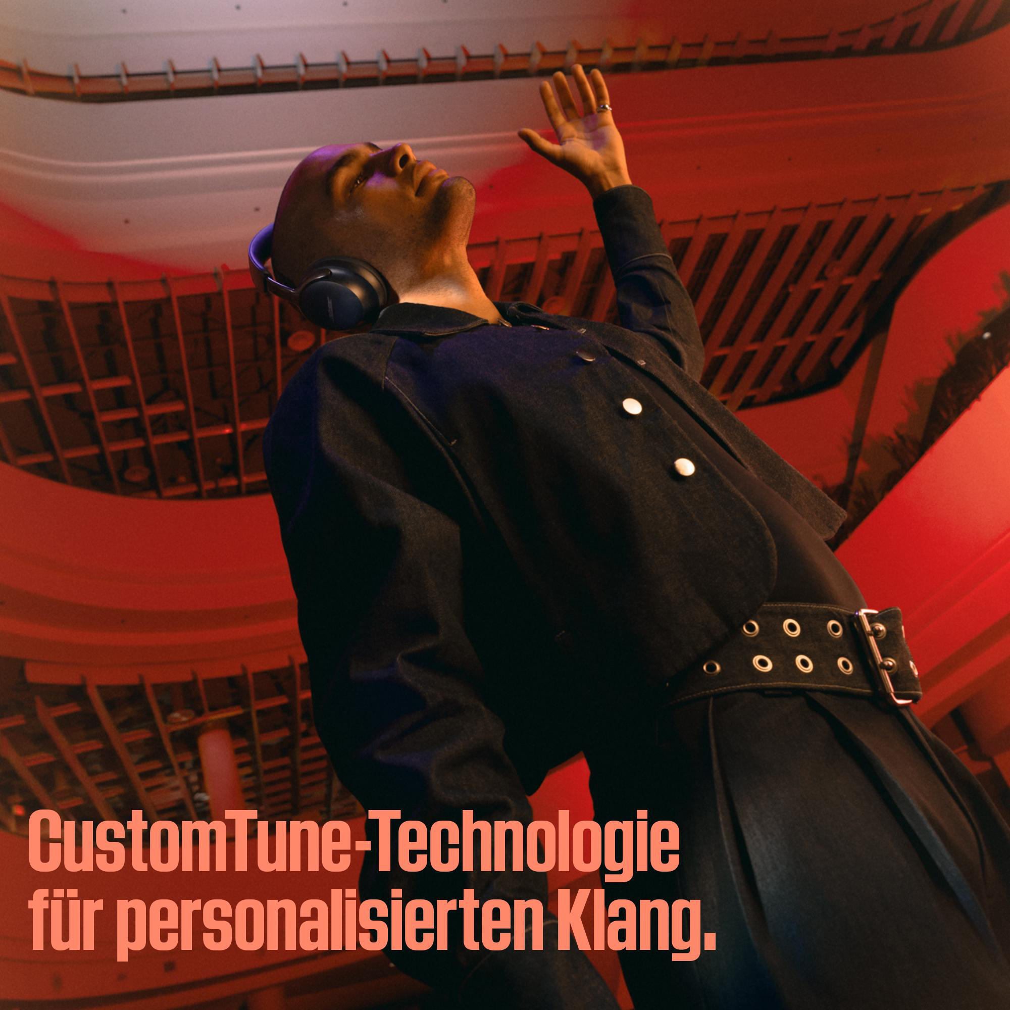 Ein Mann mit Kopfhörern und Jeansjacke blickt nach oben und hebt die Hand. Rote Beleuchtung und Text: CustomTune-Technologie.