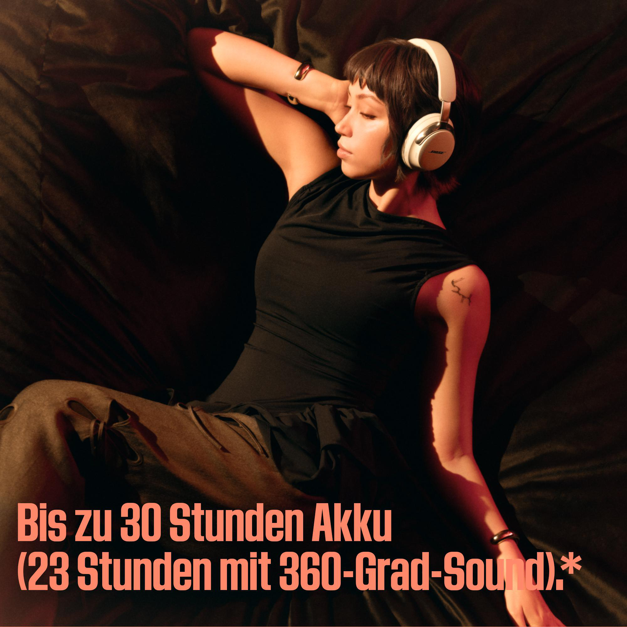 Frau mit Kopfhörern liegt auf dunkler Oberfläche. Text: Bis zu 30 Stunden Akku (23 Stunden mit 360-Grad-Sound).