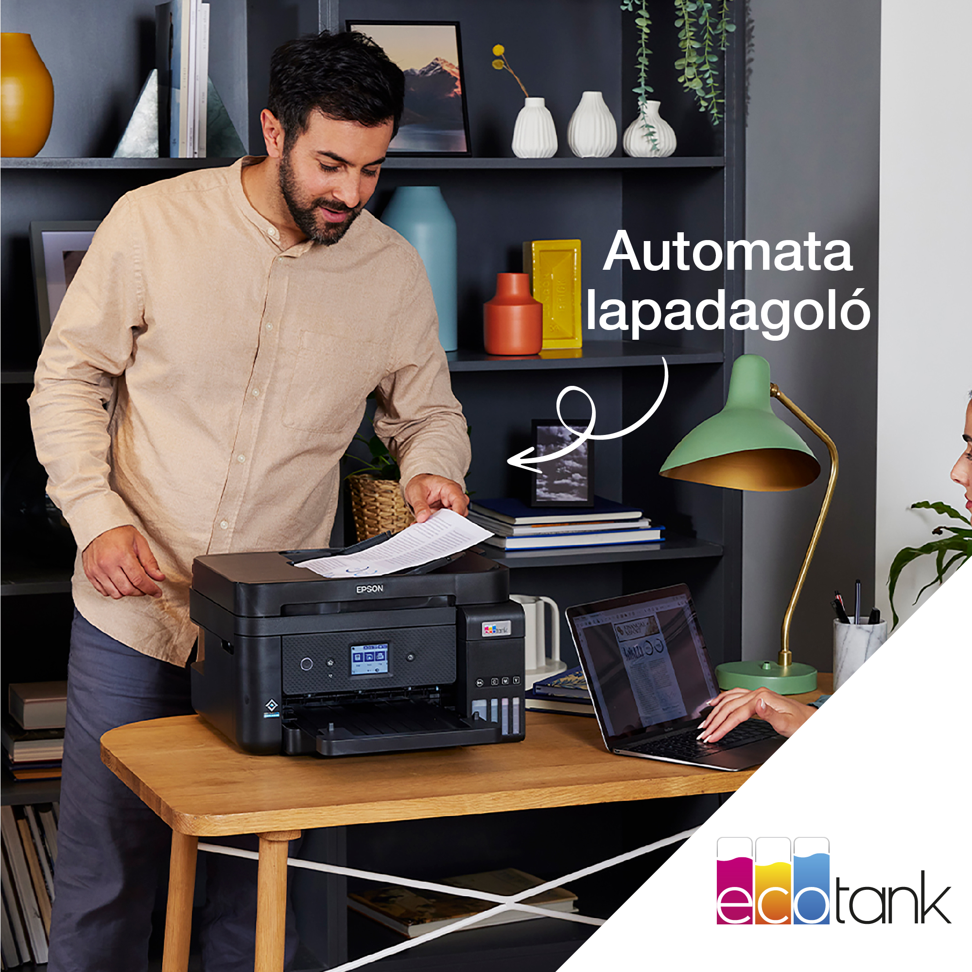 Egy férfi nyomtatót használ. Egy nő laptopot használ. Egy lámpa van az asztalon. A háttérben egy könyvespolc látható.