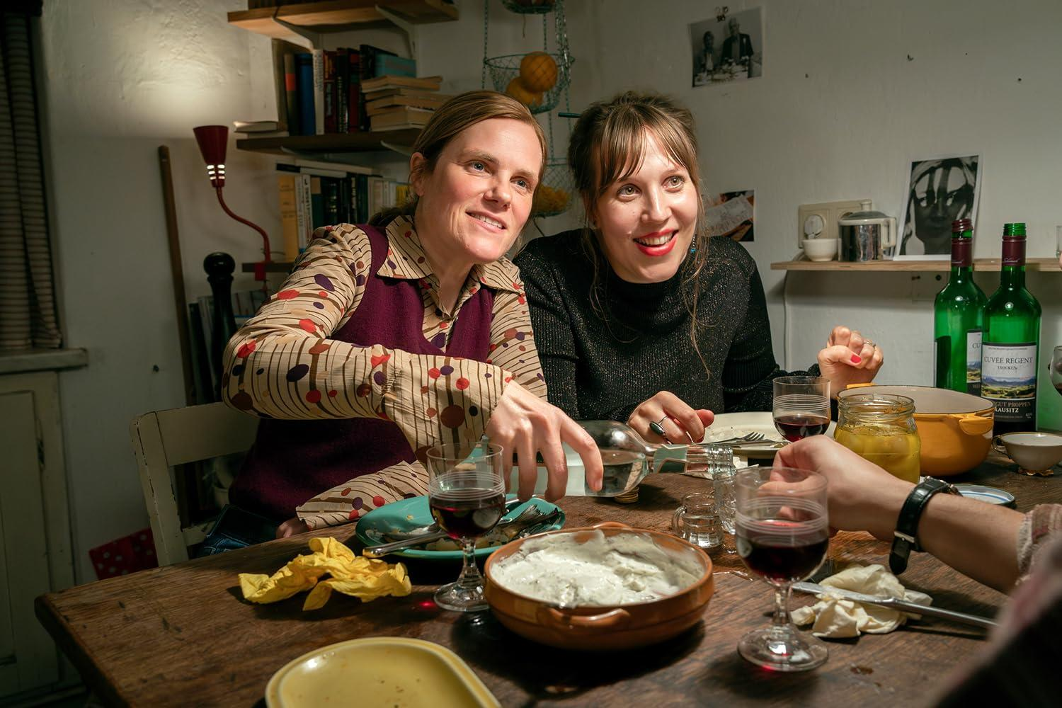Zwei Frauen an einem Tisch mit Essen und Wein. Eine gießt Wasser ein. Der Tisch ist aus Holz. Weinflaschen sind im Hintergrund.
