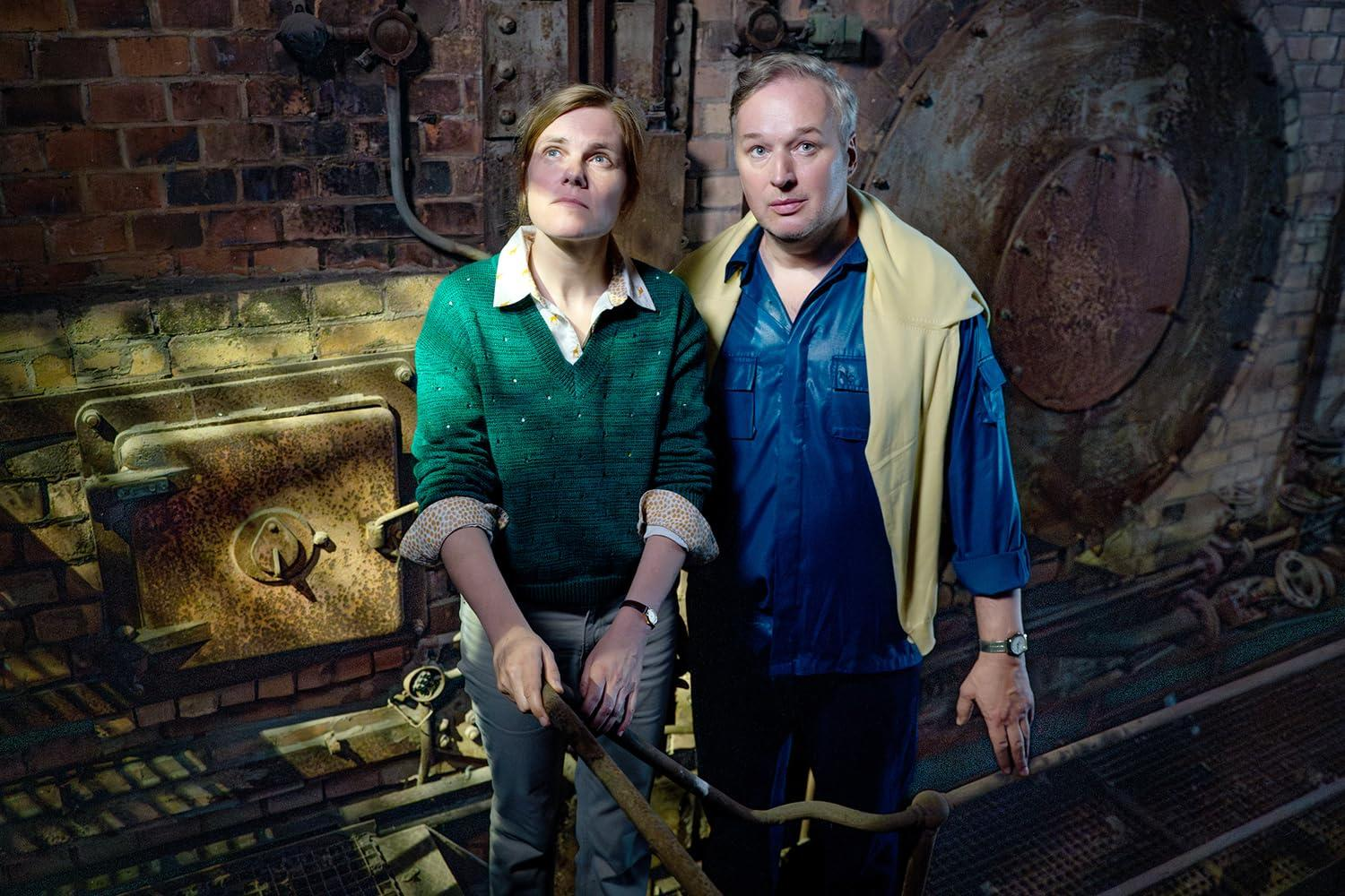 Zwei Personen stehen in einer industriellen Umgebung. Eine Frau in Grün, ein Mann in Blau. Der Hintergrund ist Ziegel und Metall.