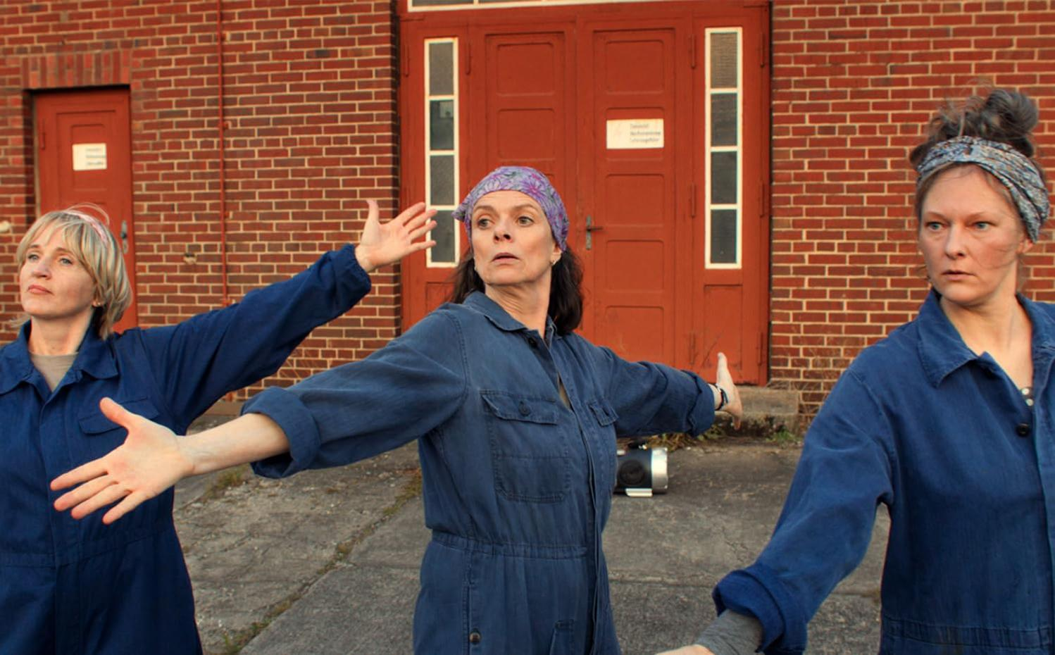 Drei Frauen in blauen Overalls stehen mit ausgestreckten Armen. Ein rotes Backsteingebäude und Türen bilden den Hintergrund.