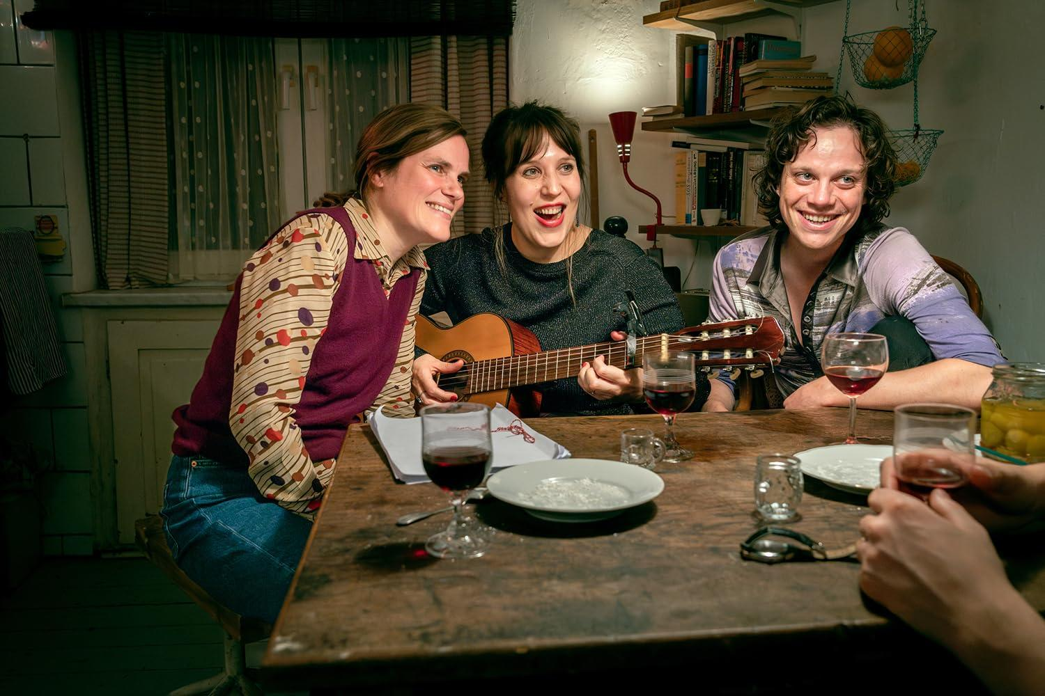 Drei Personen an einem Tisch, eine spielt Gitarre, andere lächeln und trinken Wein. Gläser, Teller und Gläser stehen auf dem Tisch.