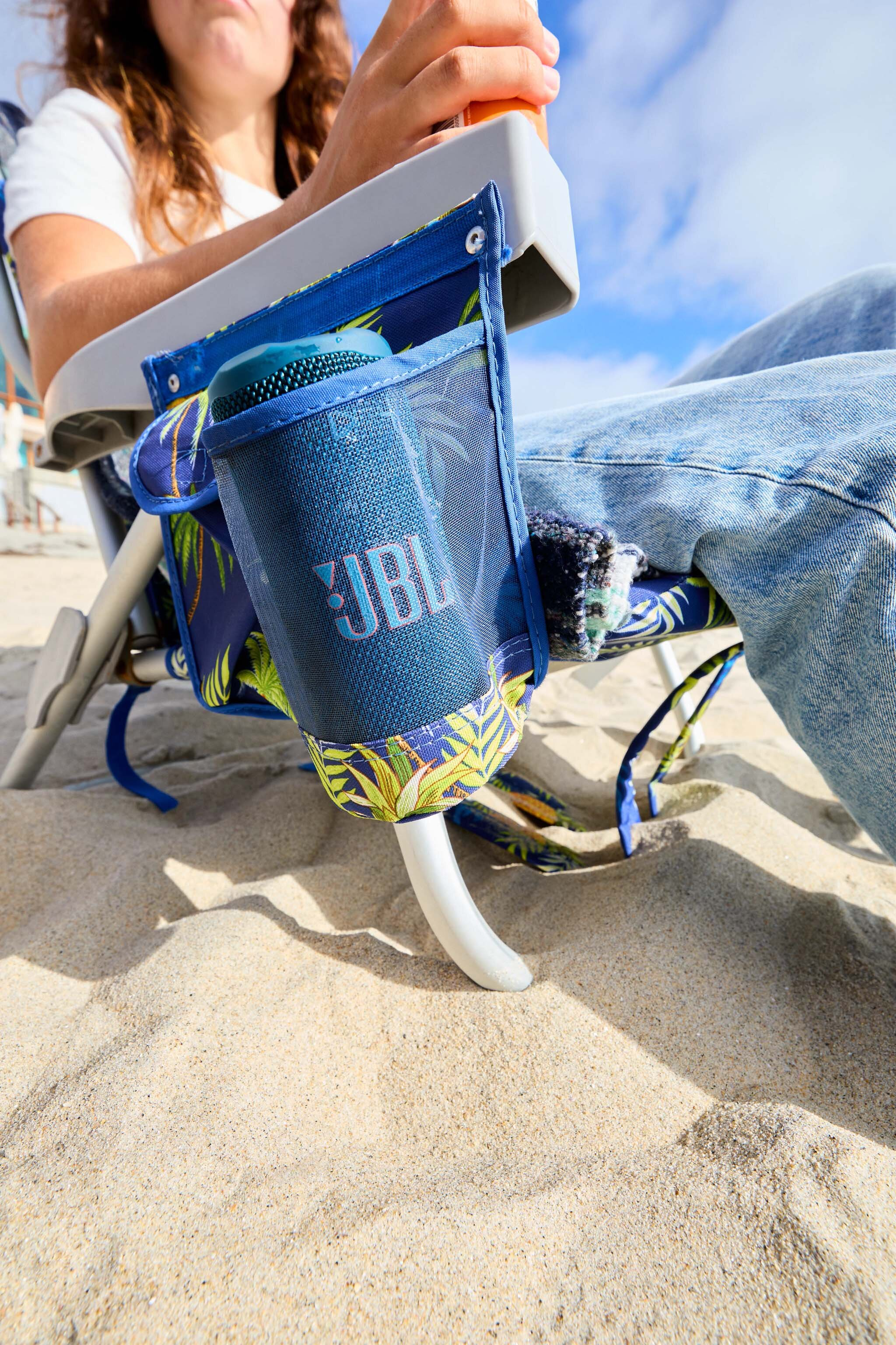 Osoba siedzi na krześle plażowym z głośnikiem JBL w bocznej kieszeni. Piasek na pierwszym planie i błękitne niebo w tle.