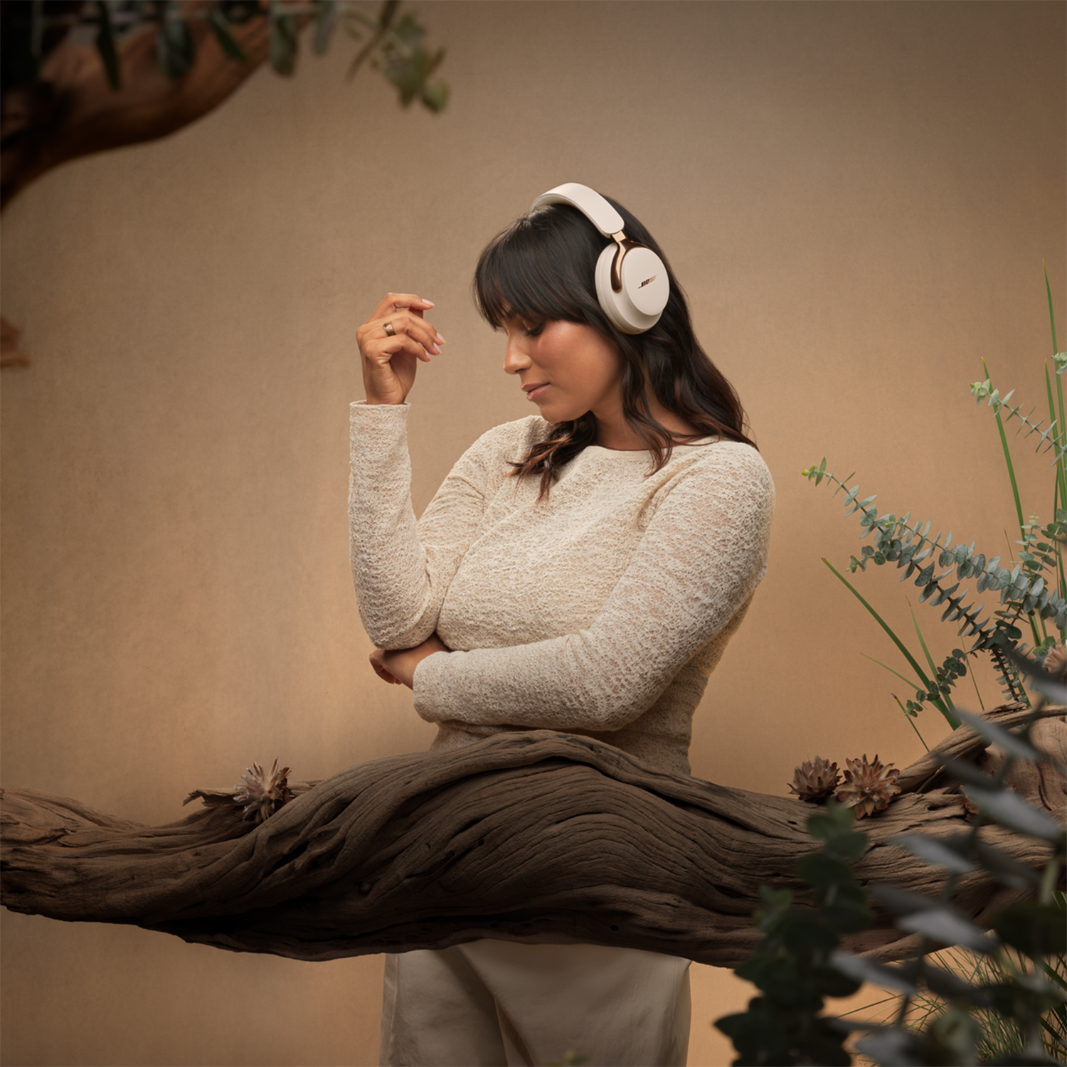 Donna con cuffie, guarda in basso, appoggiata a un ramo. Maglione beige, capelli scuri, sfondo marrone con fogliame.
