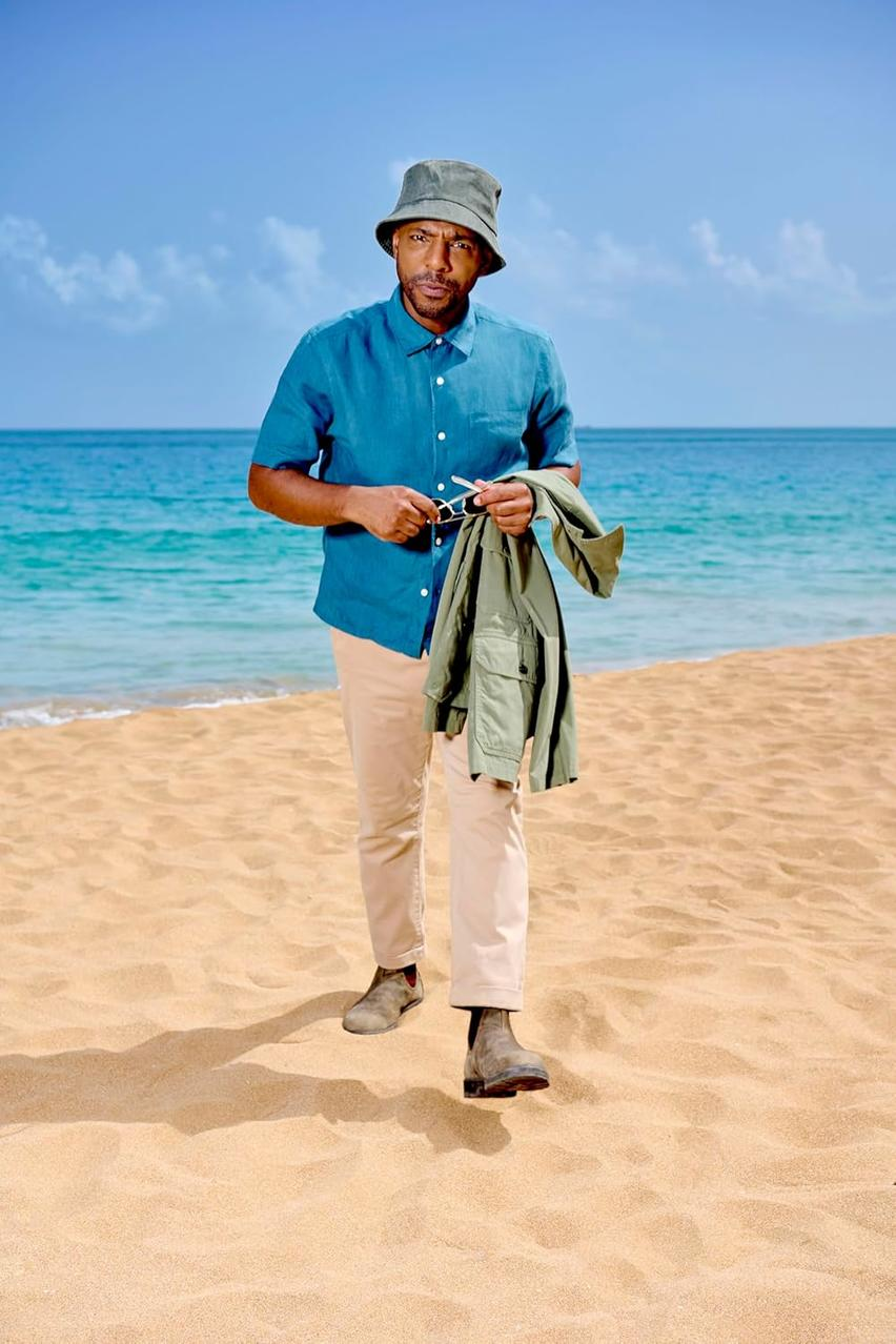 Mann am Strand mit Hut, blauem Hemd, beiger Hose und Stiefeln, hält Sonnenbrille und grüne Jacke.