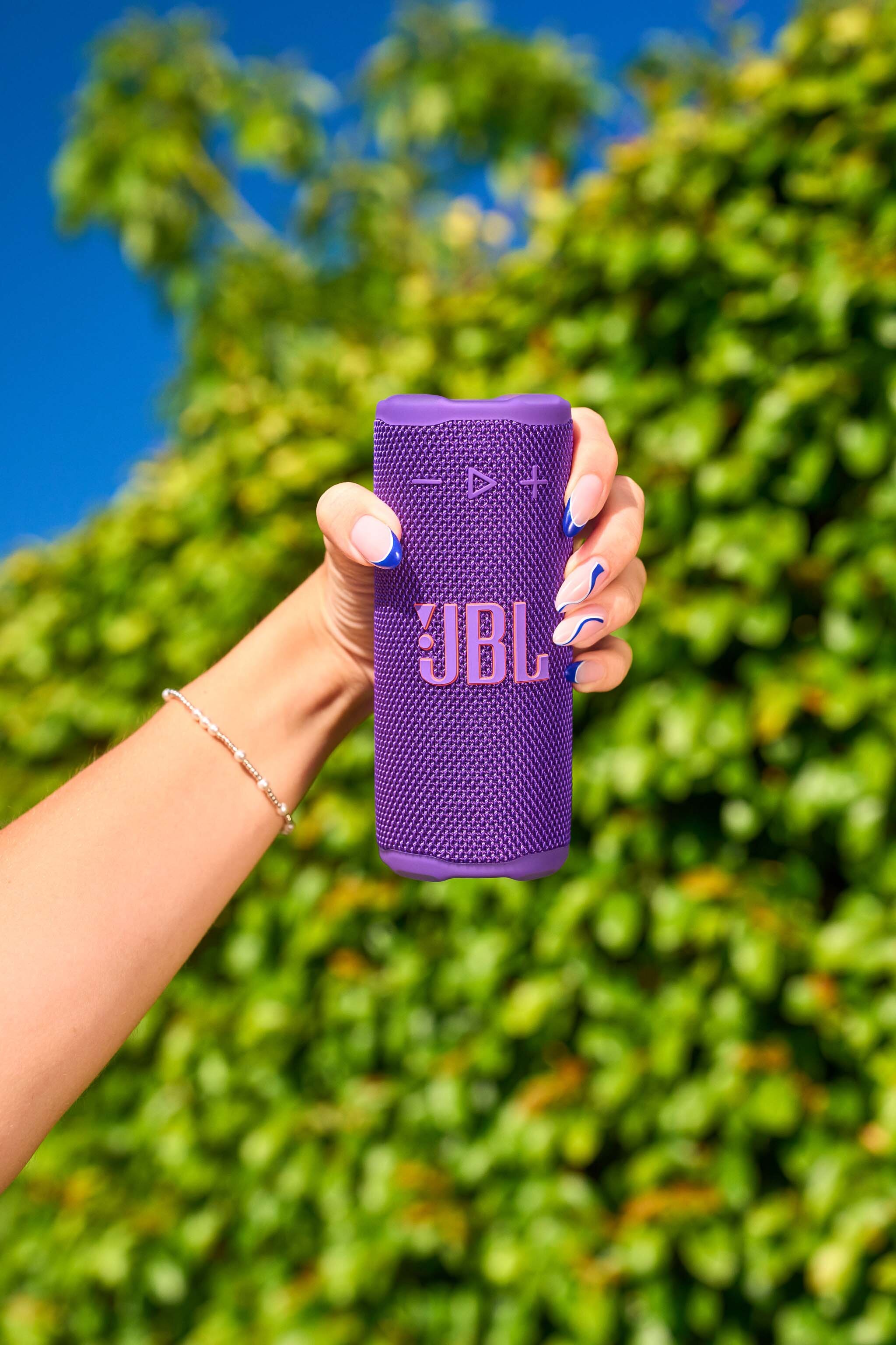 Dłoń trzyma fioletowy głośnik JBL na tle zielonych liści i błękitnego nieba.