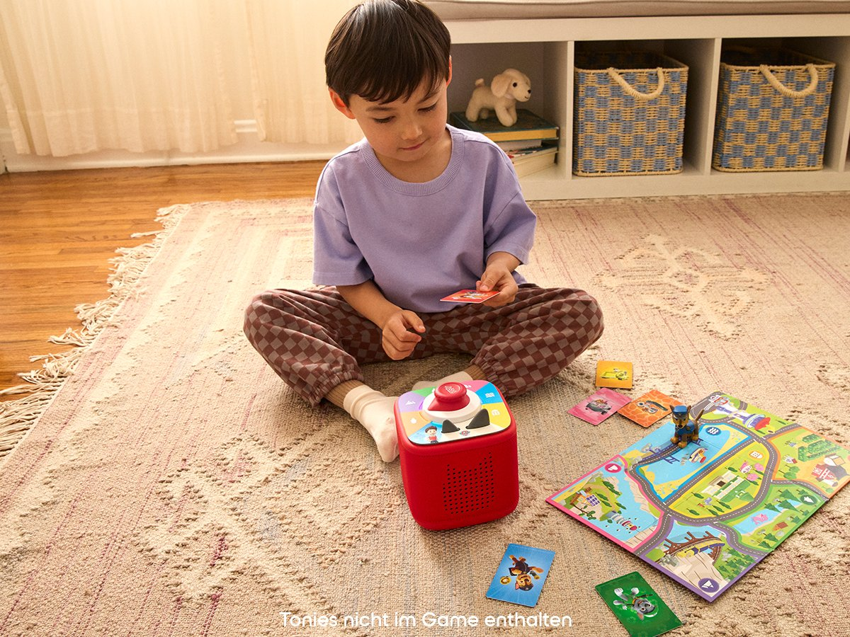 Un bambino seduto su un tappeto gioca con carte e un altoparlante rosso. Un tabellone e giocattoli sono vicini. Lo sfondo include scaffali e cestini.
