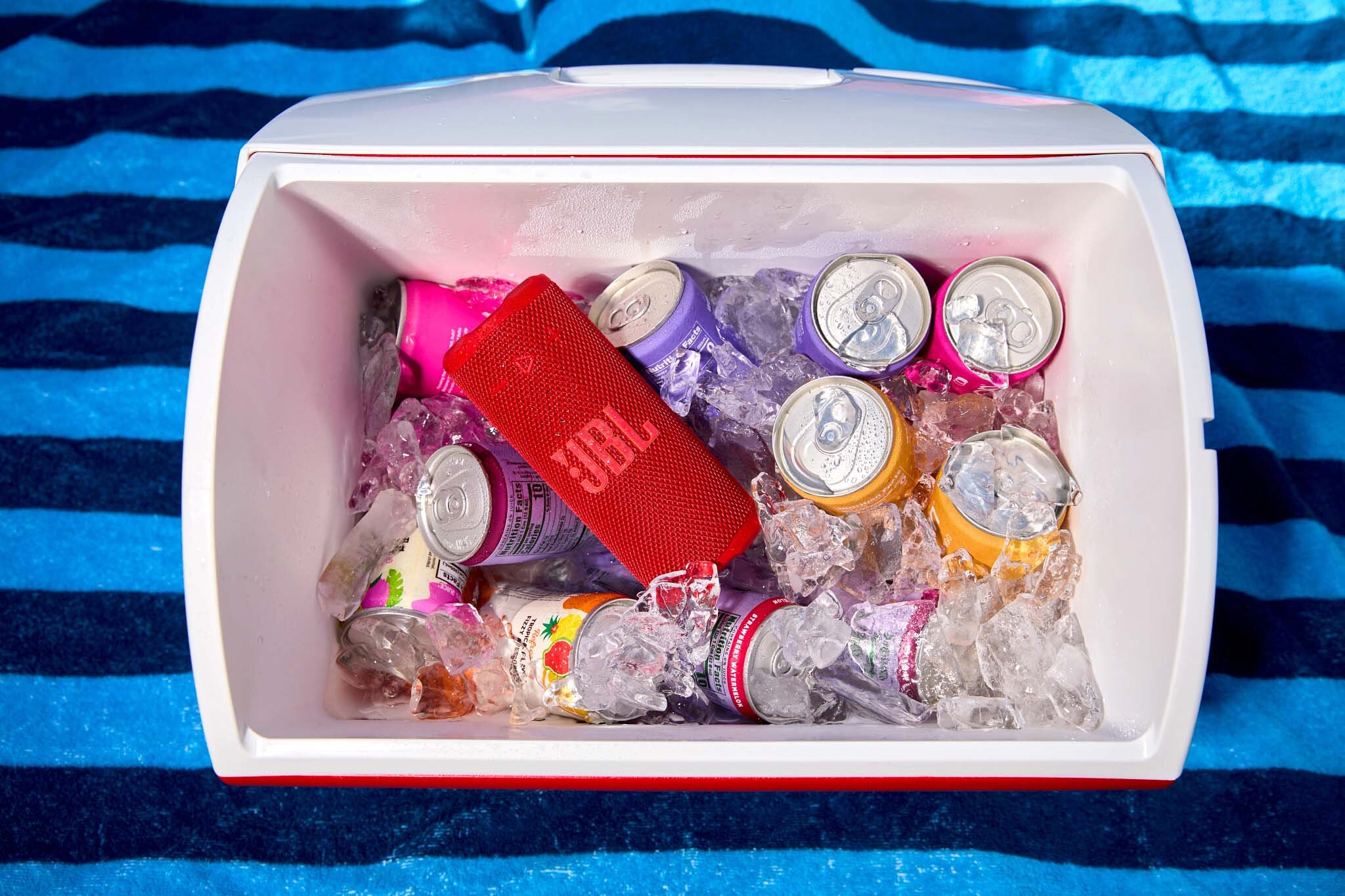 Chłodziarka wypełniona lodem, puszkami z napojami gazowanymi i czerwonym głośnikiem JBL na niebiesko-białym ręczniku w paski.