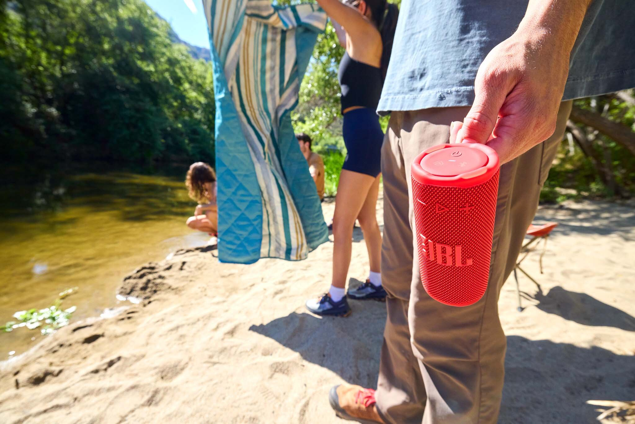 Osoba trzyma czerwony głośnik JBL na piaszczystej plaży. Ludzie rozkładają się w pobliżu rzeki. Zielone drzewa i błękitne niebo w tle.
