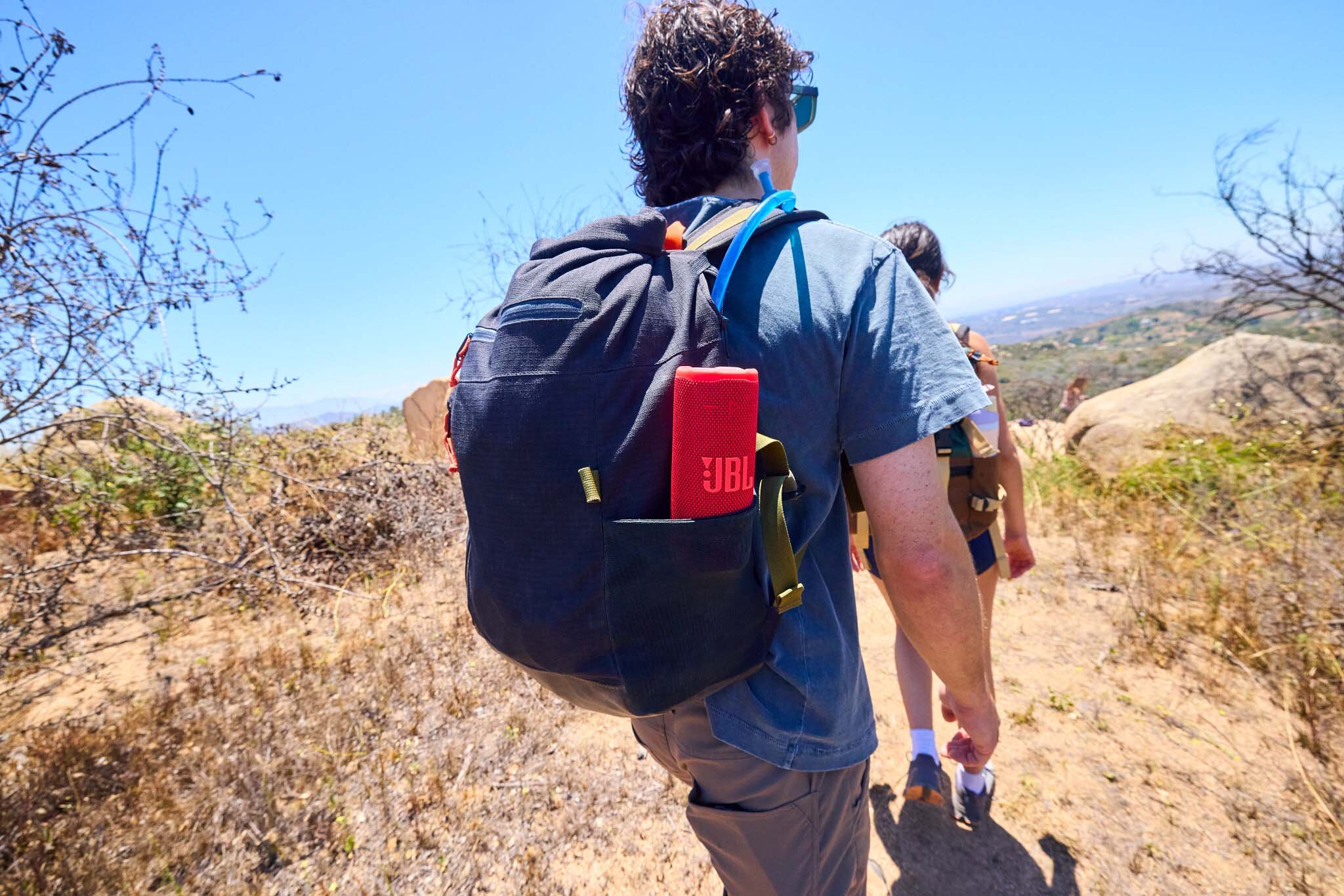 Dwie osoby wędrują w słoneczny dzień. Jeden niesie plecak z czerwonym głośnikiem JBL. Błękitne niebo i suchy krajobraz.