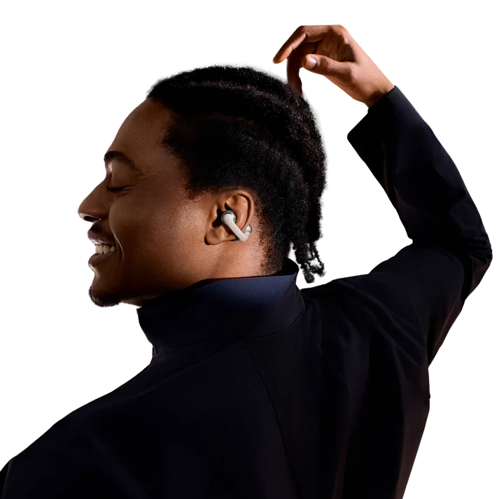 Un hombre sonriente con auriculares en la oreja, tocándose el pelo. Lleva un cuello alto azul oscuro y una chaqueta negra.