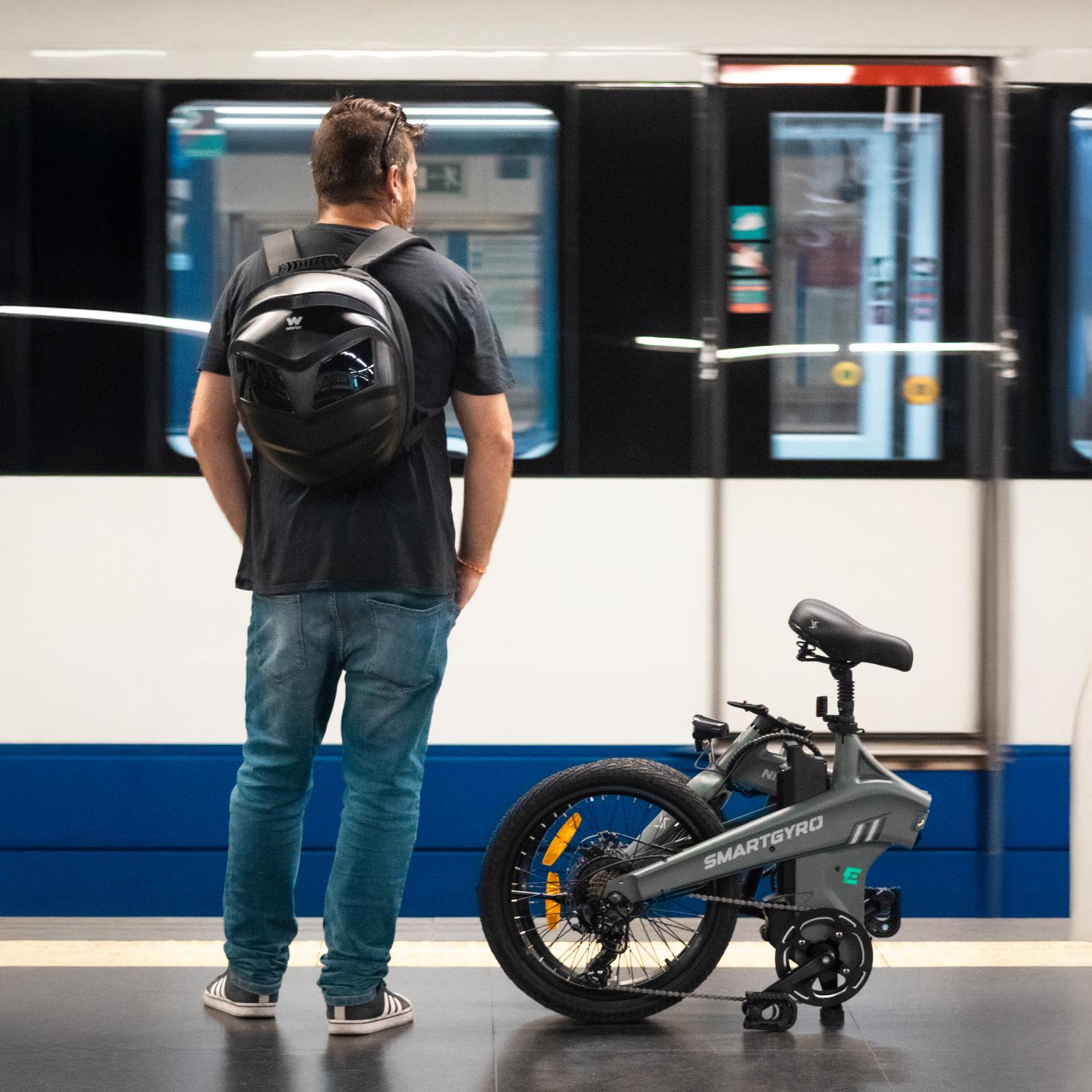 Hombre con mochila junto a una e-bike Smartgyro en estación de metro, puertas del tren abiertas al fondo.