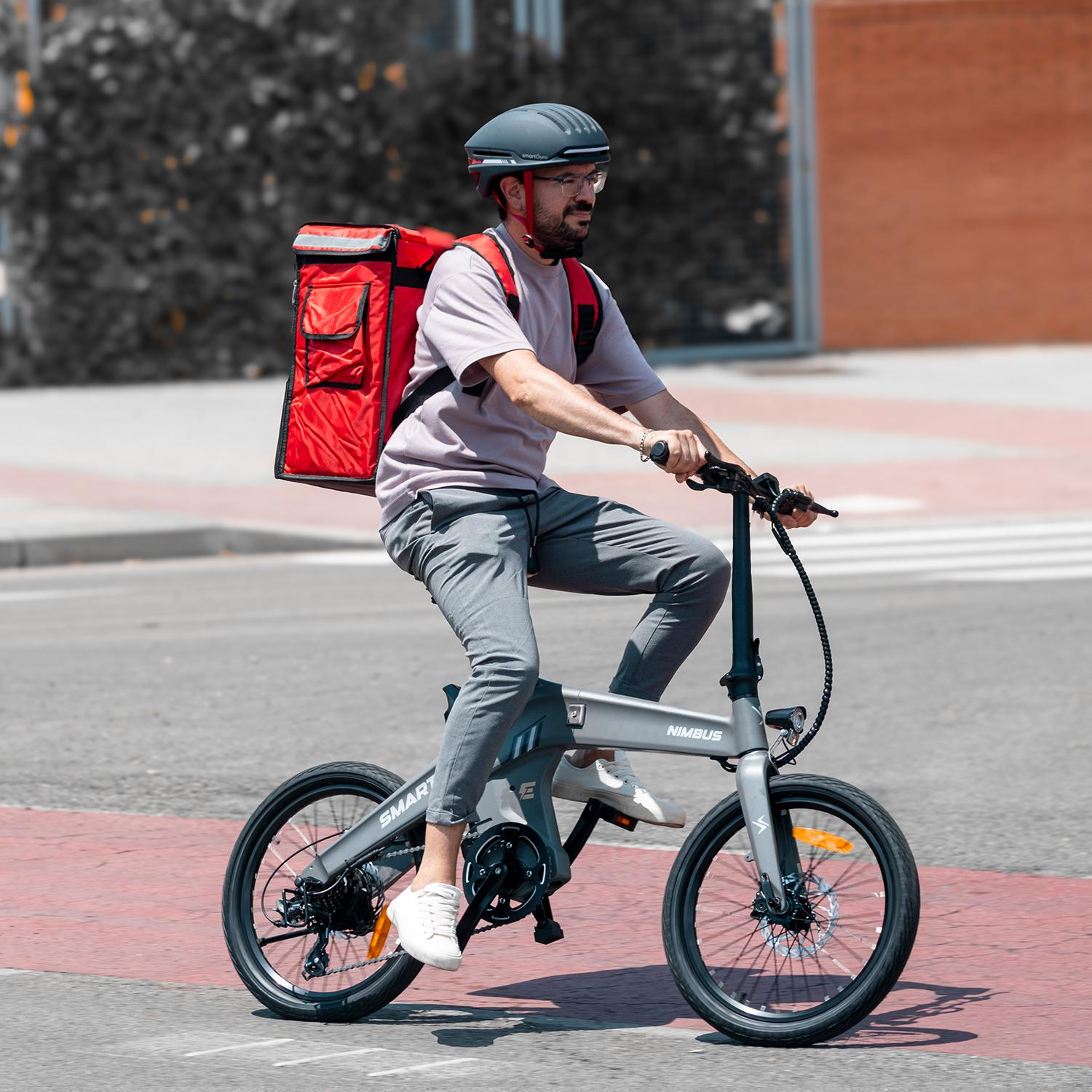Un hombre monta una e-bike gris Nimbus en la calle. Lleva casco, gafas y una bolsa de reparto roja. El fondo tiene edificios.