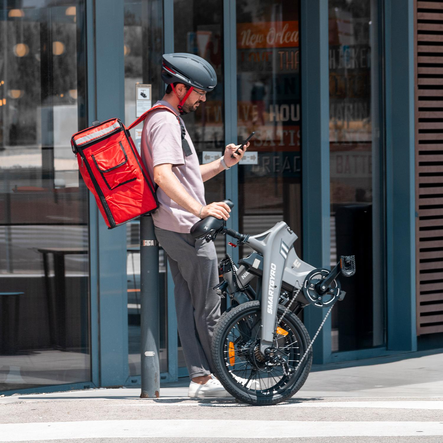 Repartidor con casco y bolsa roja, revisa el teléfono junto a una bicicleta plegable gris. Fondo: fachada de restaurante.