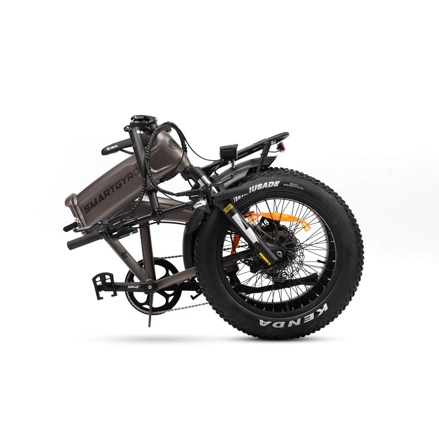 Bicicleta eléctrica gris plegable sobre fondo blanco. Cuenta con neumáticos anchos, frenos de disco y 'SMARTGYRO' en el cuadro. 'KENDA' es visible en el neumático.