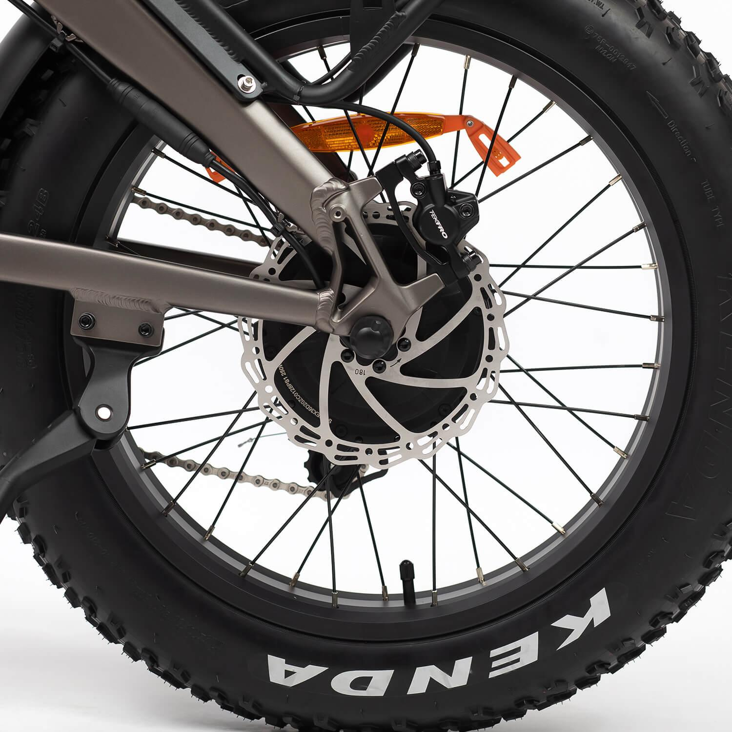 Primer plano de una rueda de bicicleta. Neumático negro con texto 'KENDA' blanco. Rotor de freno de disco plateado. Radios y cuadro en el fondo.