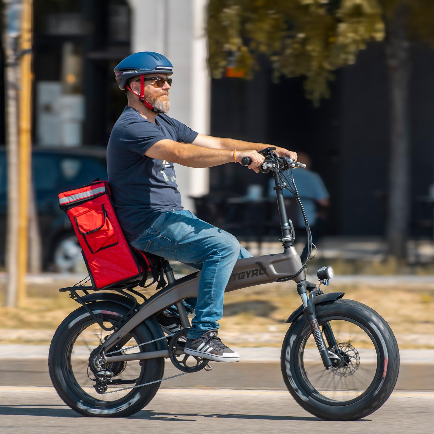 Hombre en bicicleta eléctrica con bolsa de reparto roja. Lleva casco azul y gafas de sol. La bicicleta es gris y el fondo está borroso.