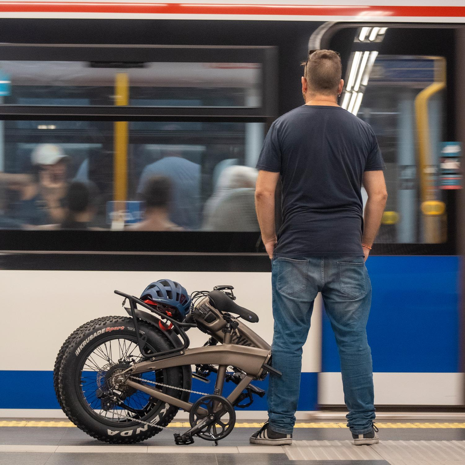 Un hombre está en un andén, mirando un tren borroso. Una bicicleta plegada con un casco está cerca. El tren está en movimiento.