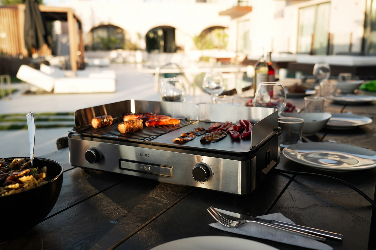 Una parrilla de acero inoxidable con salchichas y pimientos cocinados en una mesa preparada para una comida al aire libre.