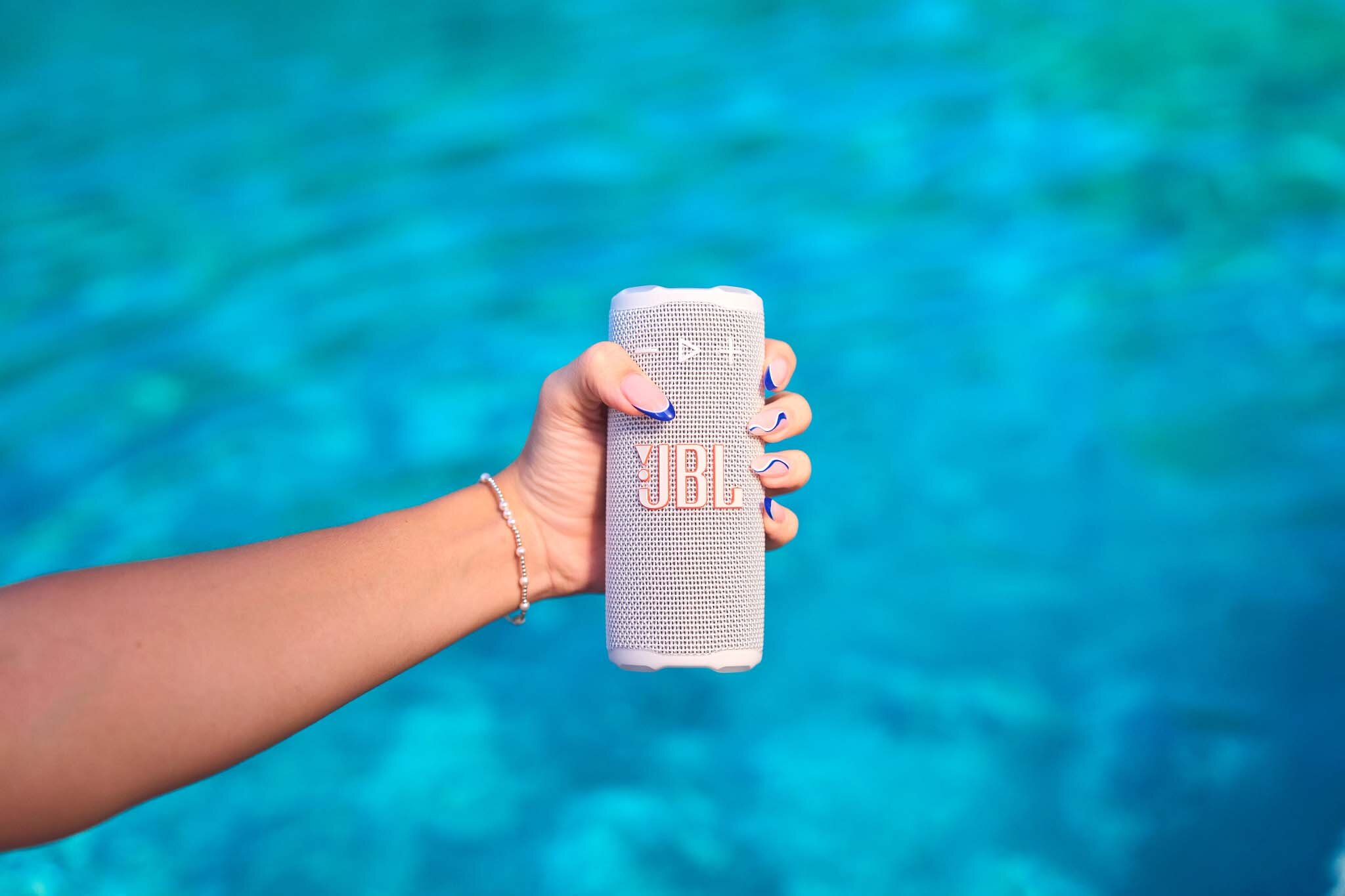 Ręka trzyma biały głośnik JBL z niebieską wodą w tle. Ręka ma niebieski lakier do paznokci.