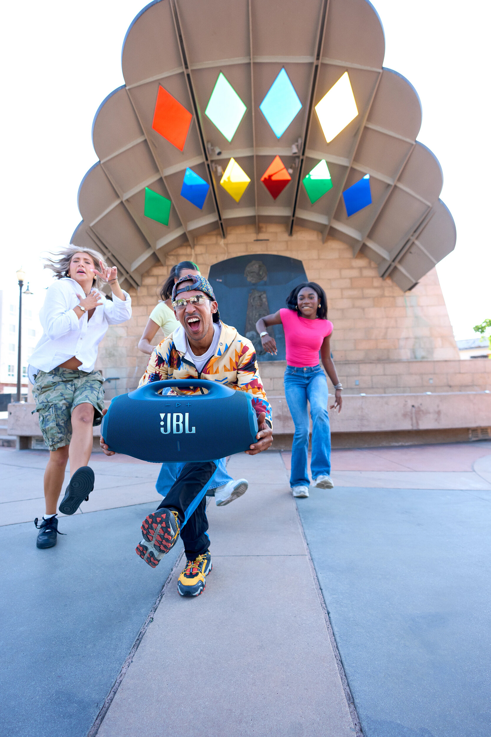 Ludzie biegają z głośnikiem JBL. Budynek z kolorowymi kształtami diamentów w tle.
