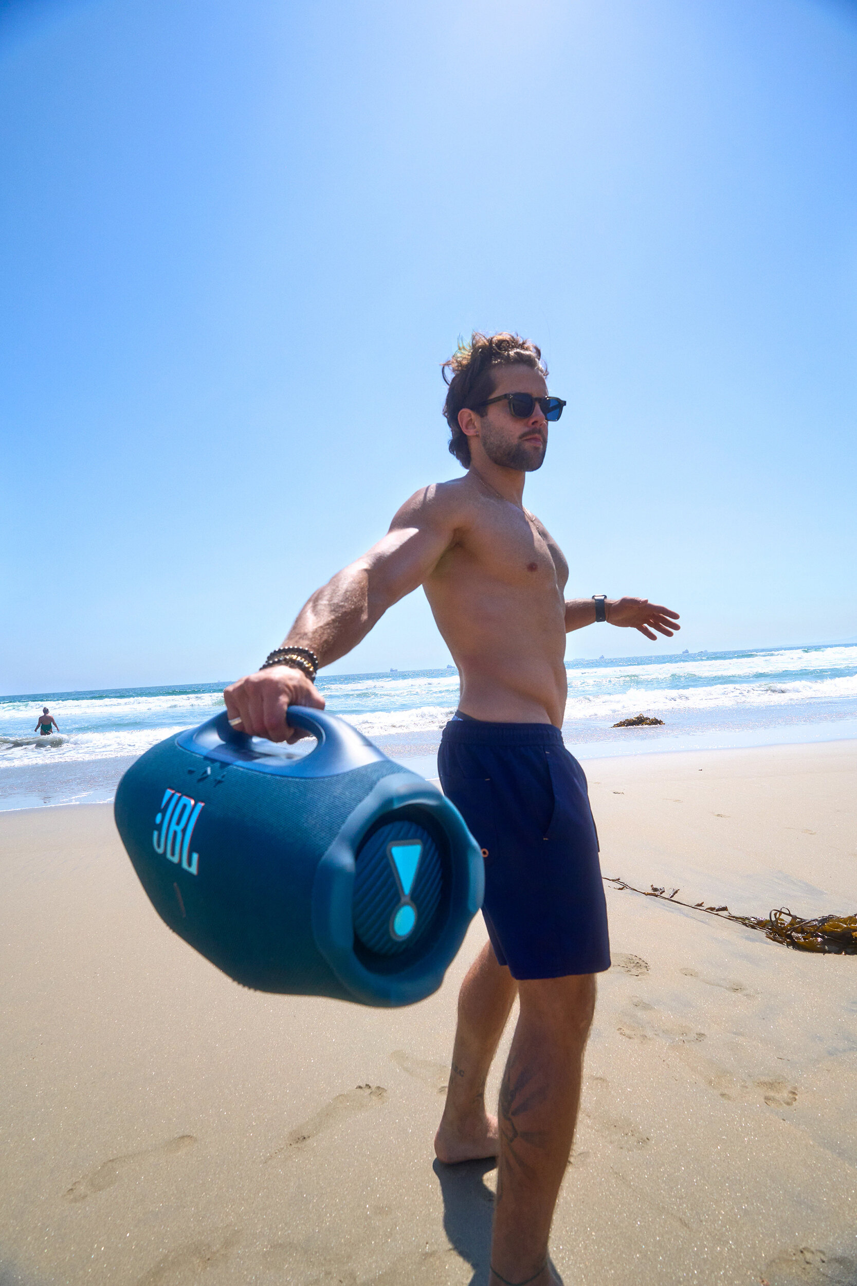 Mężczyzna na plaży trzymający niebieski głośnik JBL. Ma okulary przeciwsłoneczne i szorty. W tle ocean i niebo.